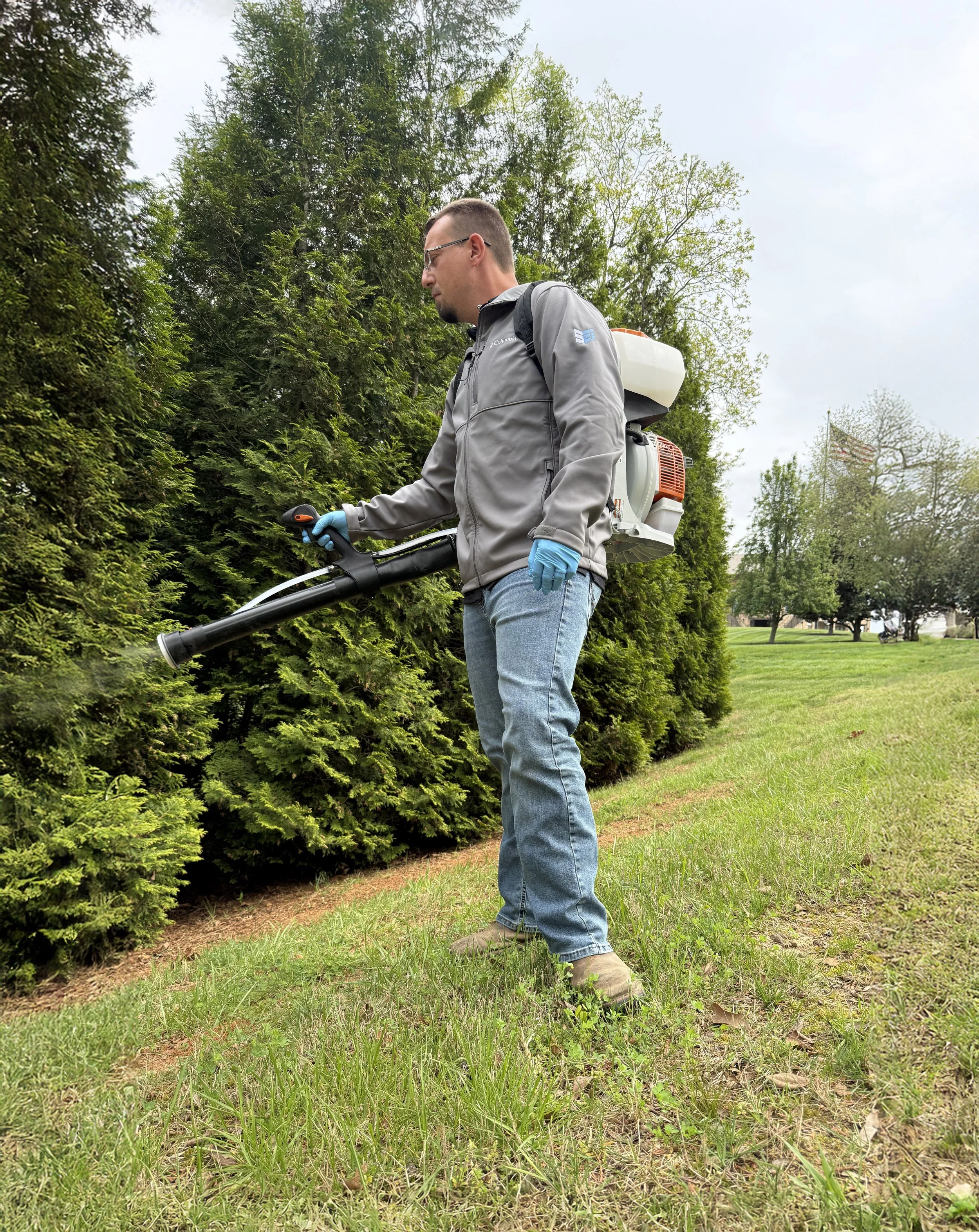 A man wearing glasses, a gray jacket, blue gloves, and jeans is using a handheld shrub trimmer to cut a hedge along a sidewalk on a cloudy day. There are trees and grass in the background.