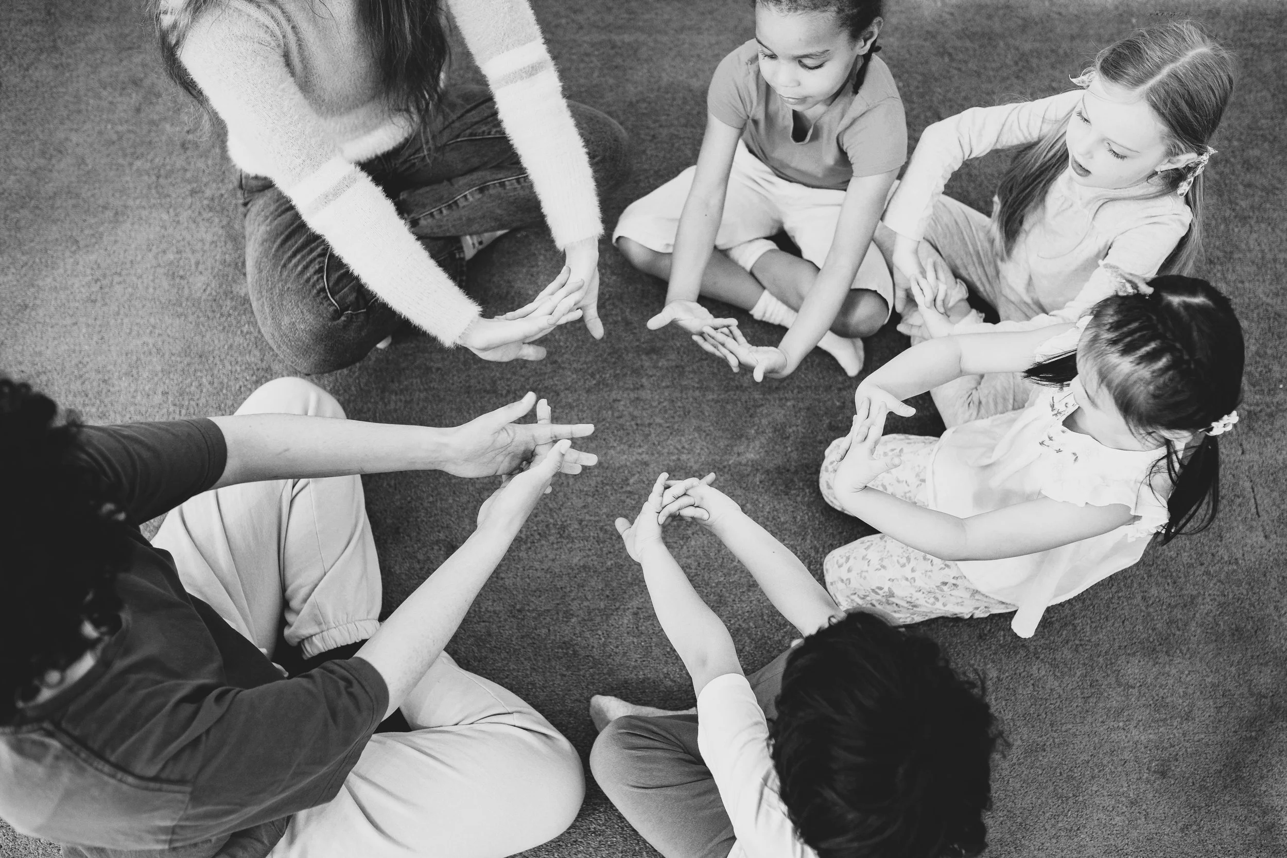 A group of children and an adult sitting in a circle on a carpeted floor, engaged in a group activity. The adult is reaching into the circle, and the children are watching and reaching with their hands.