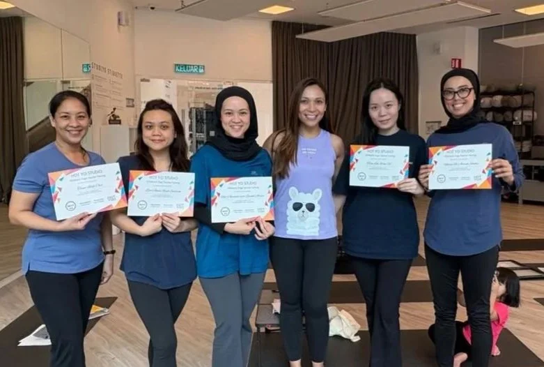 Six women standing in a row holding certificates, indoors with wooden floors and a dark curtain in the background