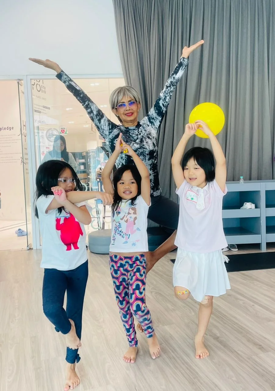 Four people, three young girls and an older woman, are playing together indoors. The woman is standing with her arms raised high, smiling, and wearing glasses. The girls are holding a yellow balloon and striking playful poses, smiling and being carefree. The environment appears to be a community center or classroom with wooden floors, a gray curtain, and some equipment in the background.