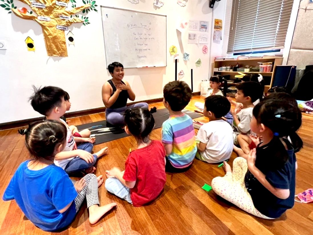 A teacher sitting on a yoga mat leading a group of young children seated on the wooden floor in a classroom. The children are facing the teacher, who is smiling and engaging with them. The classroom has educational materials, chessboard, and a whiteboard on the wall.