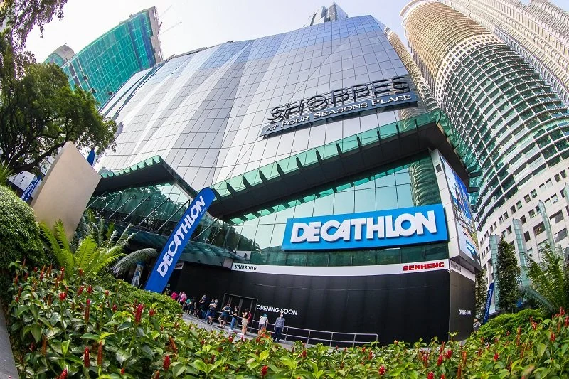 Exterior view of shops at Four Seasons Place, with Decathlon store sign and people walking near the entrance, surrounded by lush green plants.
