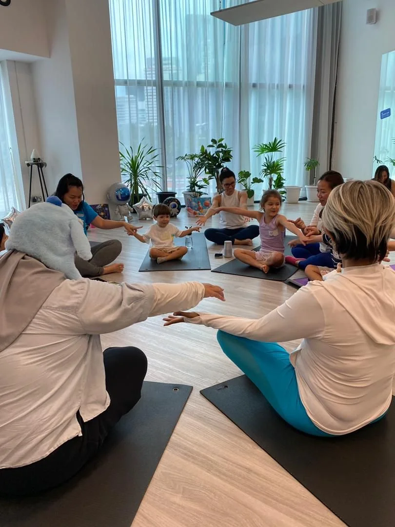 Group of children and adults sitting on yoga mats, holding hands, participating in a yoga or meditation class in a bright room with large windows and potted plants.