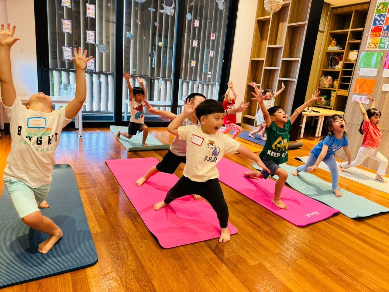 Children participating in a yoga class on colorful mats in a classroom, raising their arms and smiling.