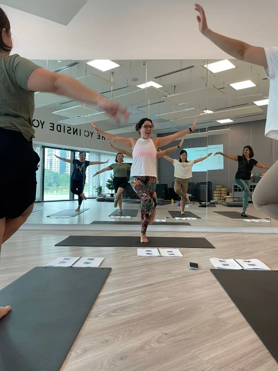Participants in a yoga class practicing tree pose in front of a large mirror, with instructor smiling in the center, mats and open books on the floor.