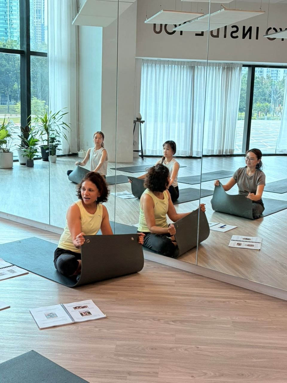 Four women practicing yoga or meditation in a bright studio with large windows, plants, and a mirrored wall, seated on yoga mats with open books and papers in front of them.