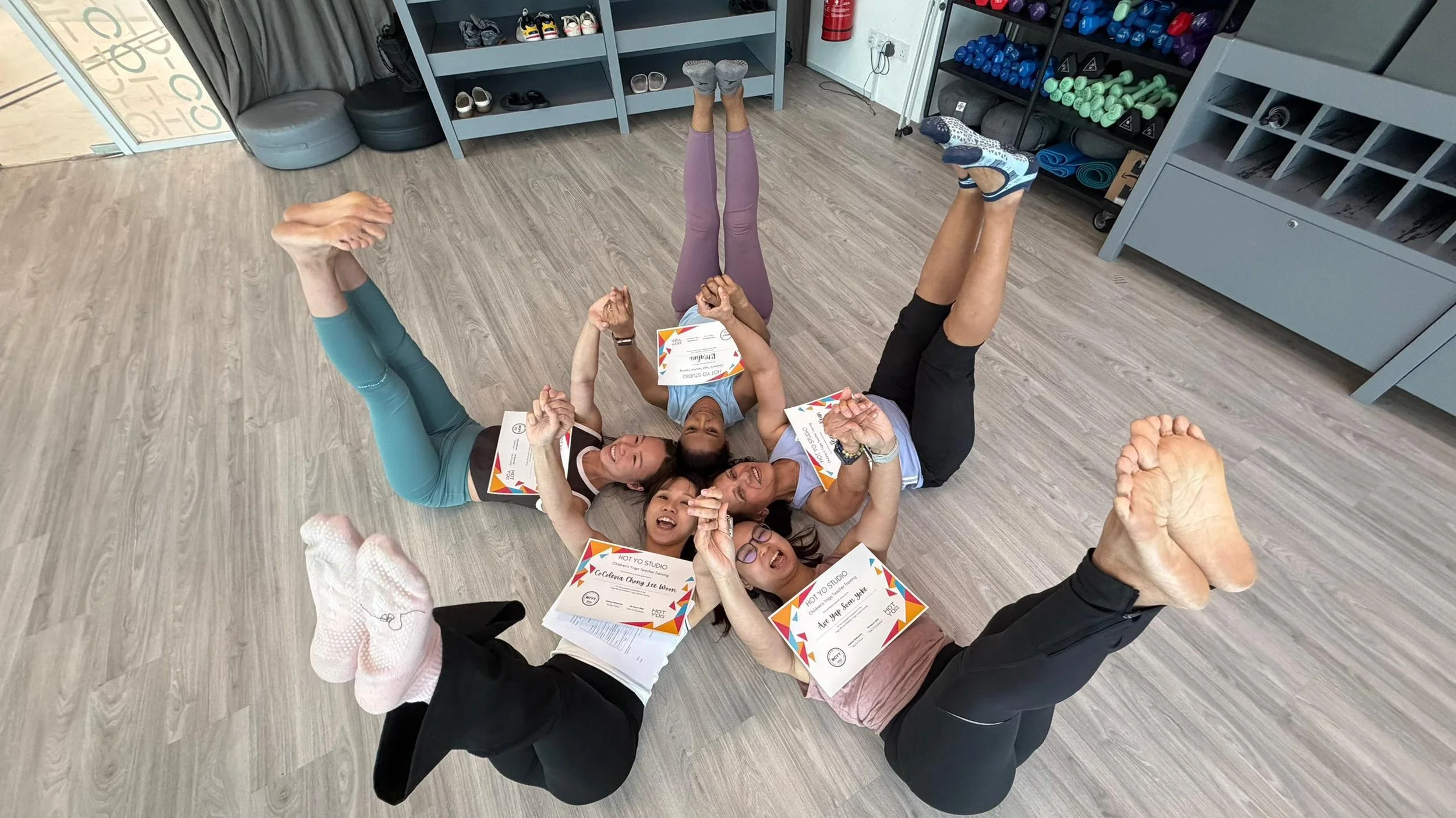 Group of six women lying on the wooden floor in a circle, holding hands and smiling. They are wearing athletic clothing and have certificates in front of them. Exercise equipment and shelves are visible in the background.