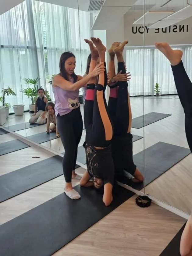 A yoga class in a bright studio with large windows and plants. An instructor assists a student with a headstand pose by holding her legs. Other students sit on mats in the background.