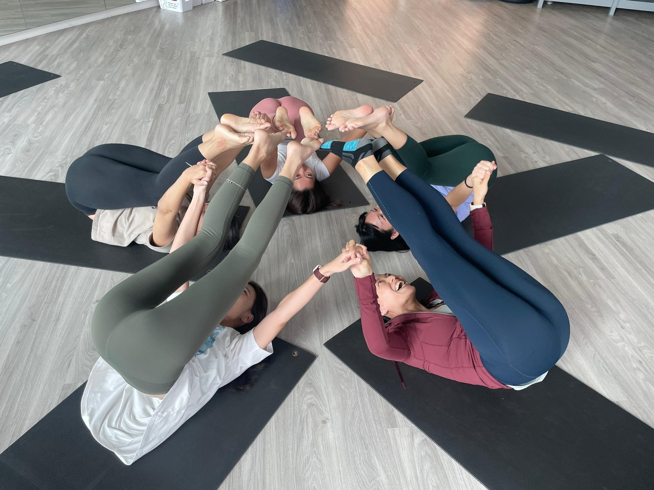 Group of people doing a yoga pose together in a studio with black mats on a wooden floor.