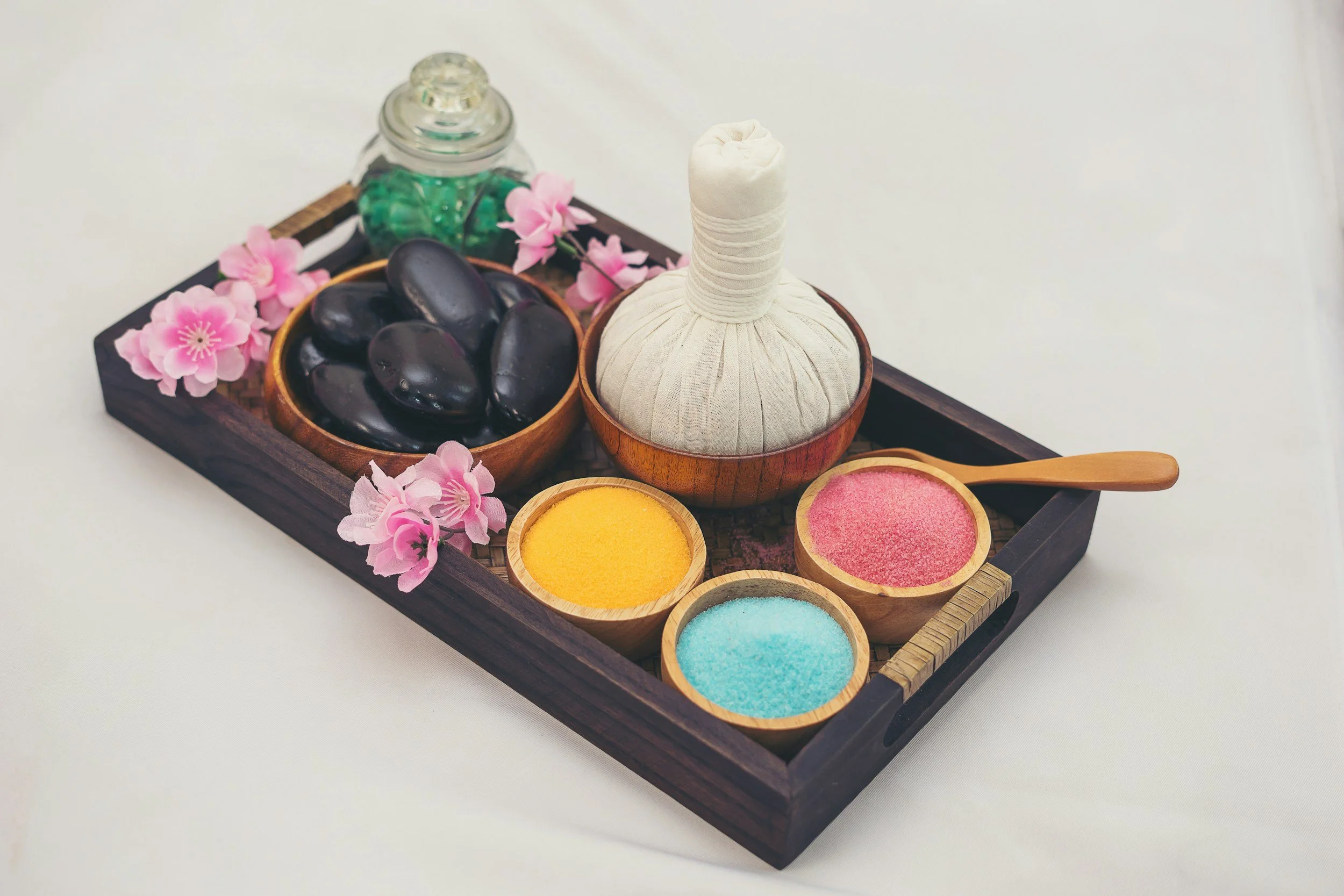 A wooden tray with pink flowers, black stones, a small glass jar with green contents, a white fabric-wrapped herbal compress ball, and four small bowls of colorful powders and textures on a white surface.