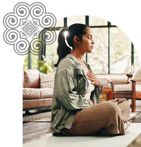 Woman meditating in a cozy, sunlit living room, sitting cross-legged on a yoga mat with her eyes closed and hand on her chest.