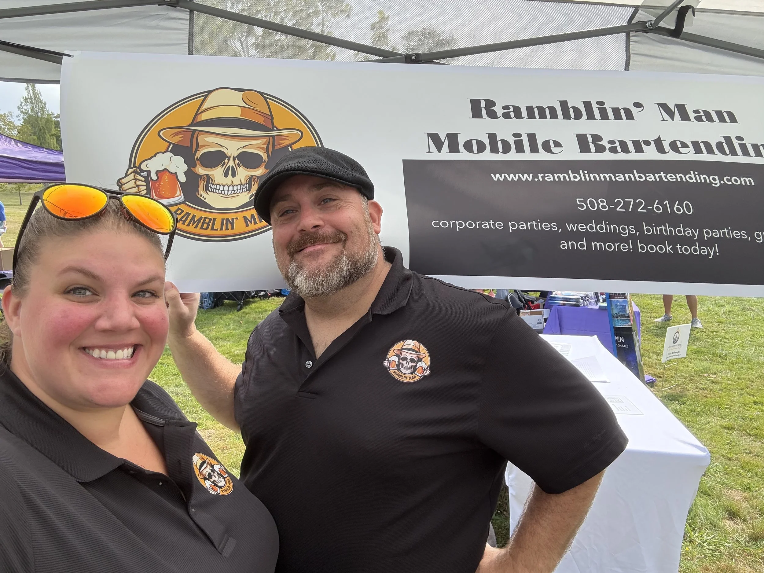 Close-up of a smiling woman's face with sunglasses on her head and a man next to her, both wearing black shirts with Ramblin' Man logo, standing in front of a banner for Ramblin' Man Mobile Bartending at an outdoor event.