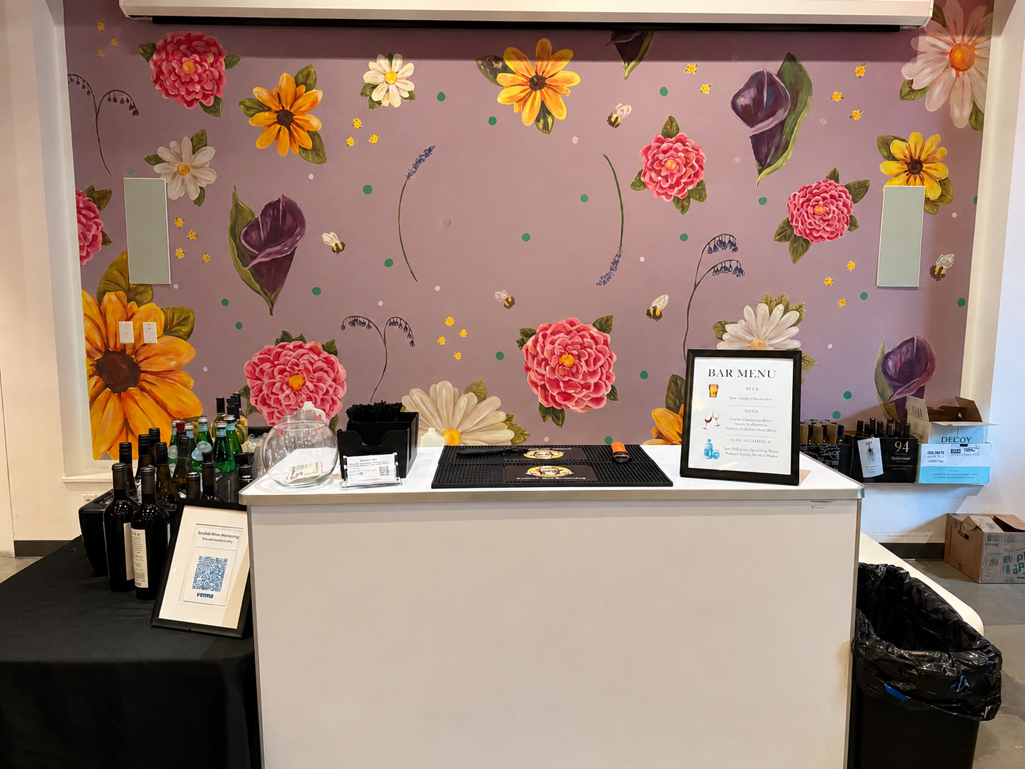 Bar setup with a floral pink and purple wall, bottles of beer and non-alcoholic beverages, a framed bar menu, and a black countertop with bar tools.
