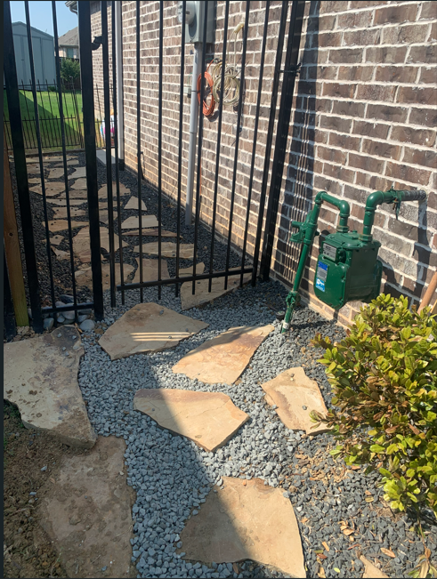 Outdoor pathway with stepping stones, gravel, a green gas meter, and a black metal gate along a brick wall.