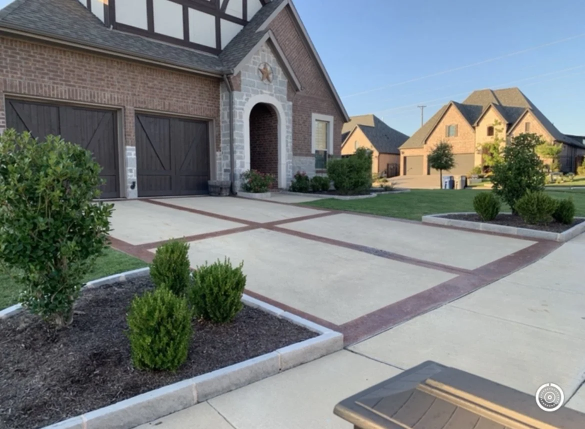 A suburban house with a brick and stone exterior, a dark wood garage door, and a driveway with red brick border patterns. There are small landscaped garden beds with bushes and plants, and other houses are visible in the background under a clear blue sky.