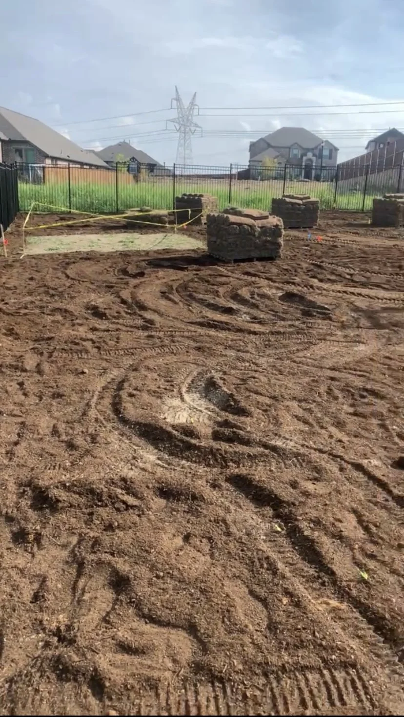 A construction site with disturbed earth, tire marks, and two brick structures, surrounded by a black metal fence, with houses and a power line tower in the background.