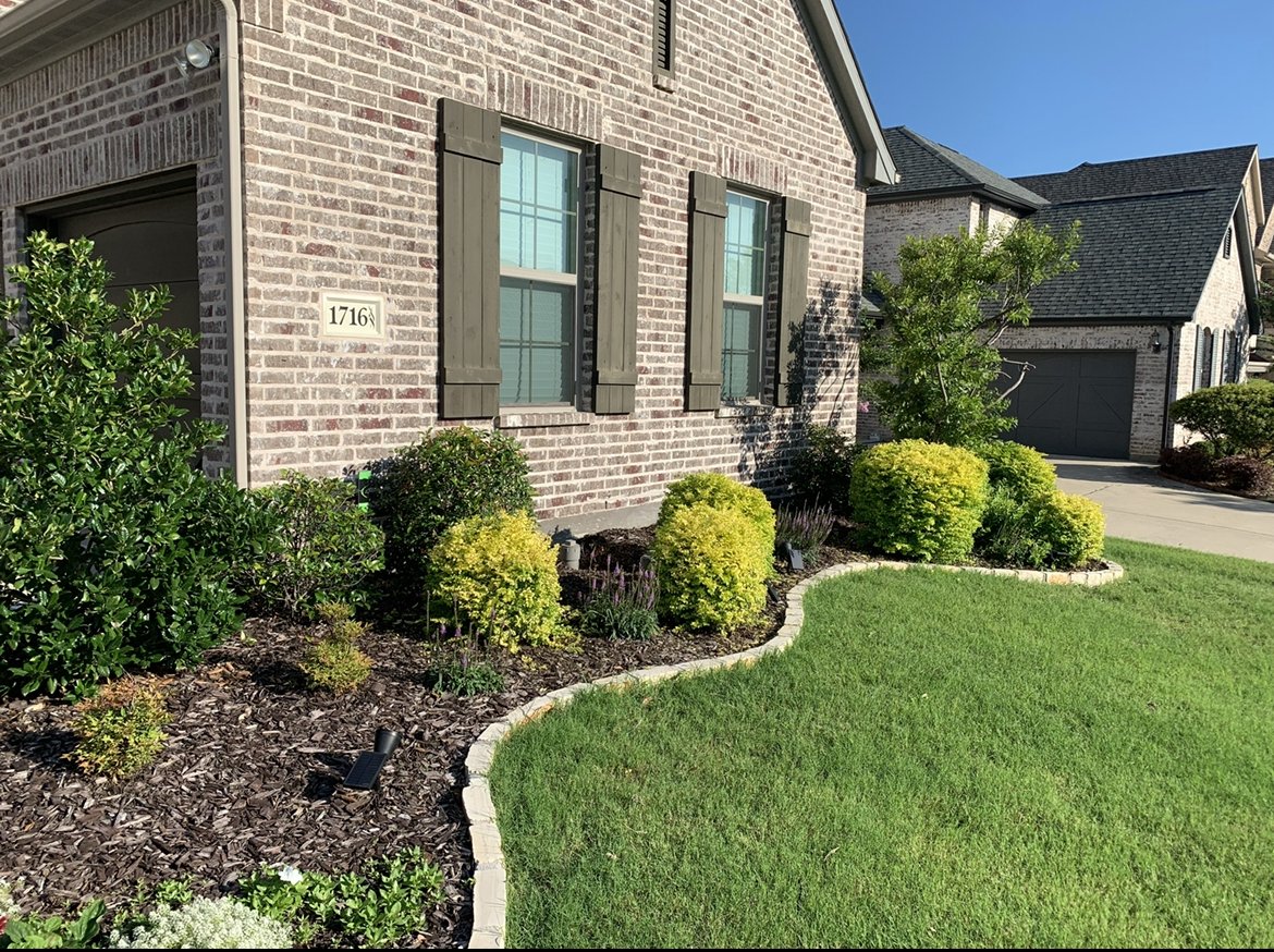 Front yard of a brick house with green bushes, yellow shrubs, a small tree, and a manicured lawn. The house has dark shutters, a house number 1716, and a garage door visible in the background.