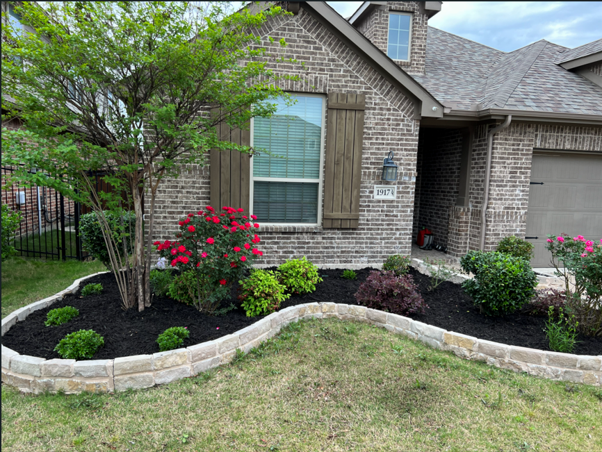 Front yard garden with a tree, colorful flowers, and green bushes, bordered by a curved stone wall, in front of a brick house with window shutters.