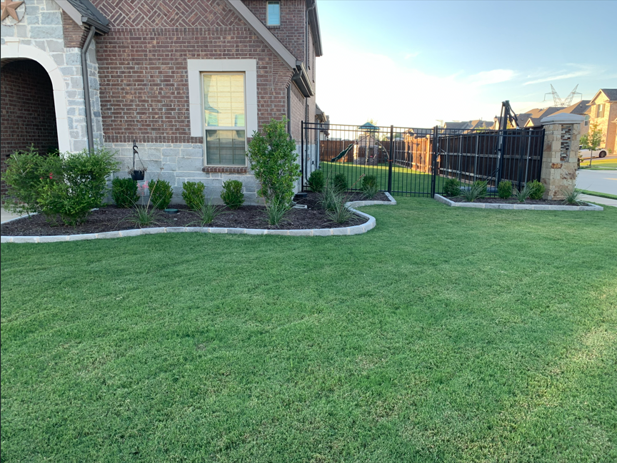 A landscaped backyard with a well-maintained lawn, small bushes, and a brick house with a closed window. A fenced gate with a playset inside is visible in the background. The yard is outlined with light-colored bricks, and there is a stone wall built