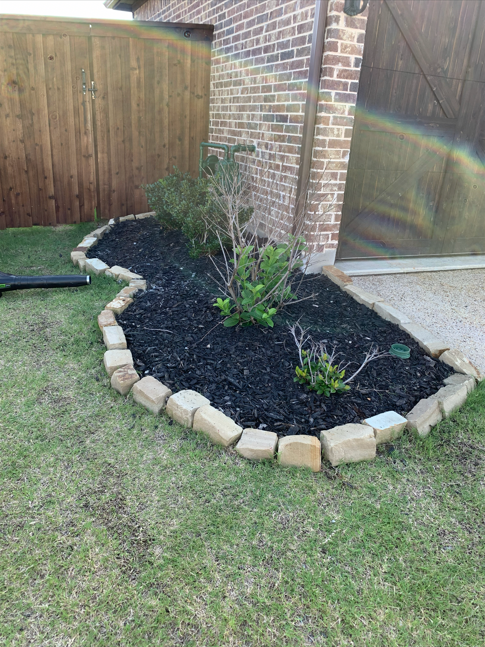 A landscaped garden bed bordered with light-colored bricks, containing small plants and bushes, planted in black mulch, next to a brick wall and a wooden fence.