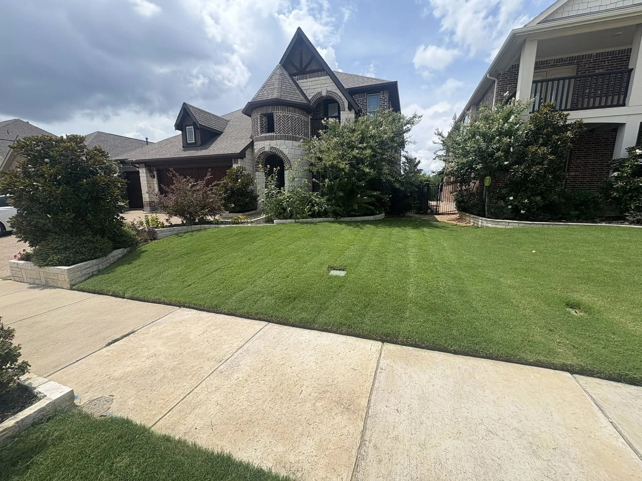 Front yard with well-manicured green lawn, stone-bordered garden beds, surrounding bushes, and a large house with stone and brick exterior, peaked roofs, and a balcony on the right side.