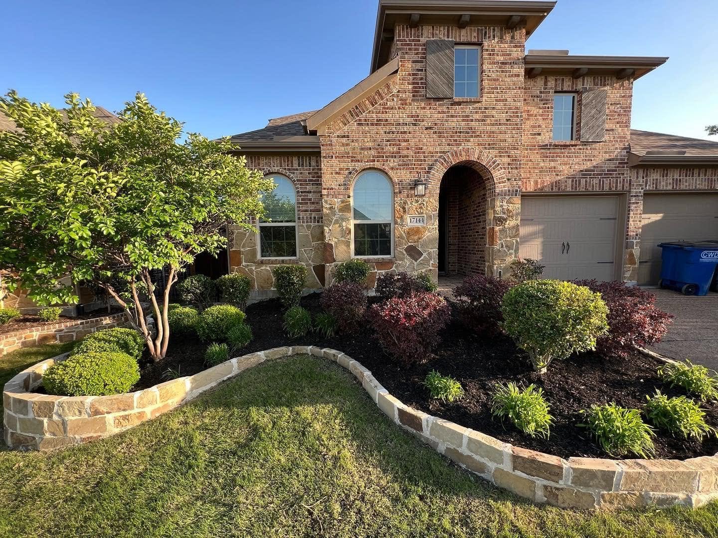 Front view of a brick house with a landscaped garden featuring bushes, a small tree, and a stone border.