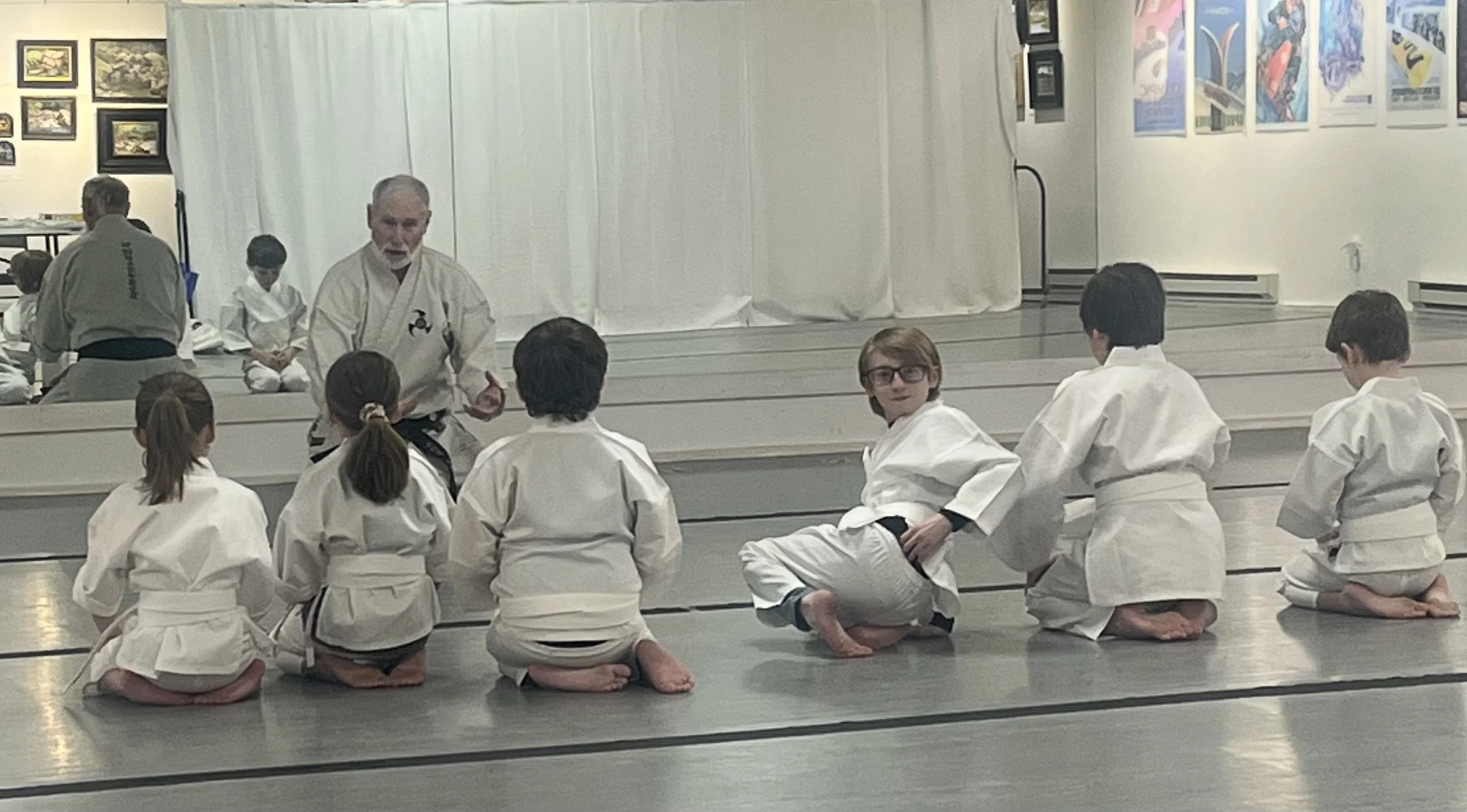 A group of children and an instructor in a martial arts class, all dressed in white gis, sitting in a kneeling position on a dojo mat, listening to the instructor. The classroom has framed pictures on the wall and a curtain in the background.