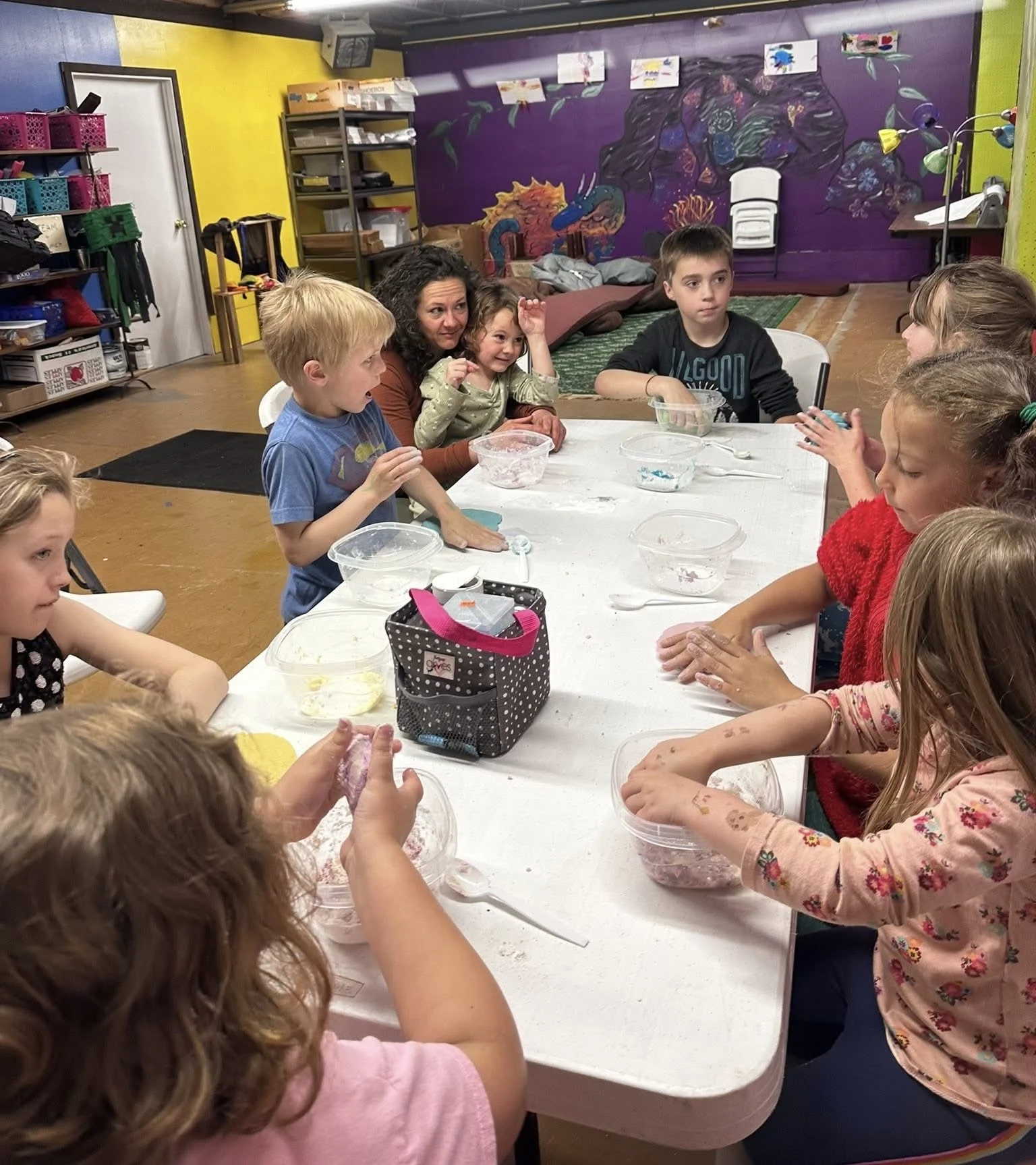 Children and an adult sitting around a white table engaging in a craft activity with bowls of colored materials inside a vibrant classroom, decorated with murals and shelves filled with supplies.