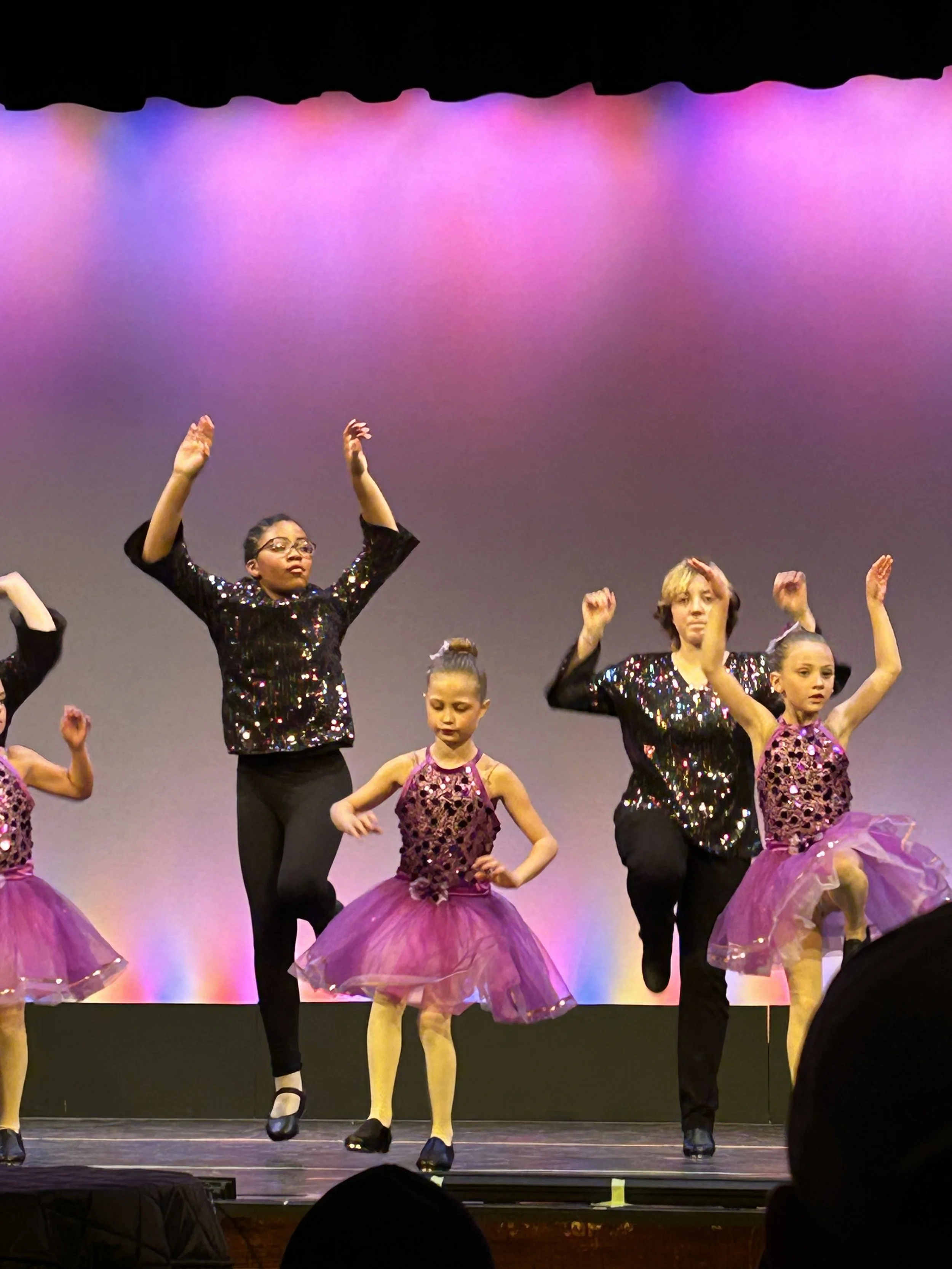 Group of young dancers performing on stage, wearing colorful costumes and black dance shoes, against a vibrant, colorful background.