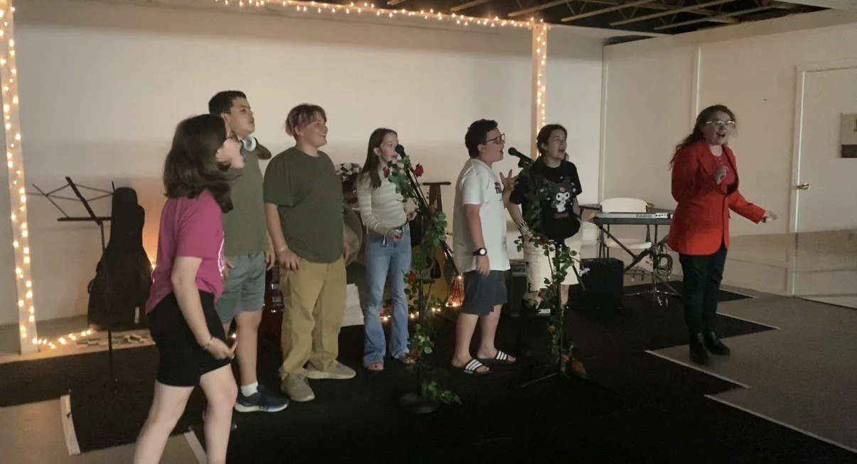 A group of seven young people singing karaoke on a small stage decorated with string lights and greenery, with musical instruments in the background.