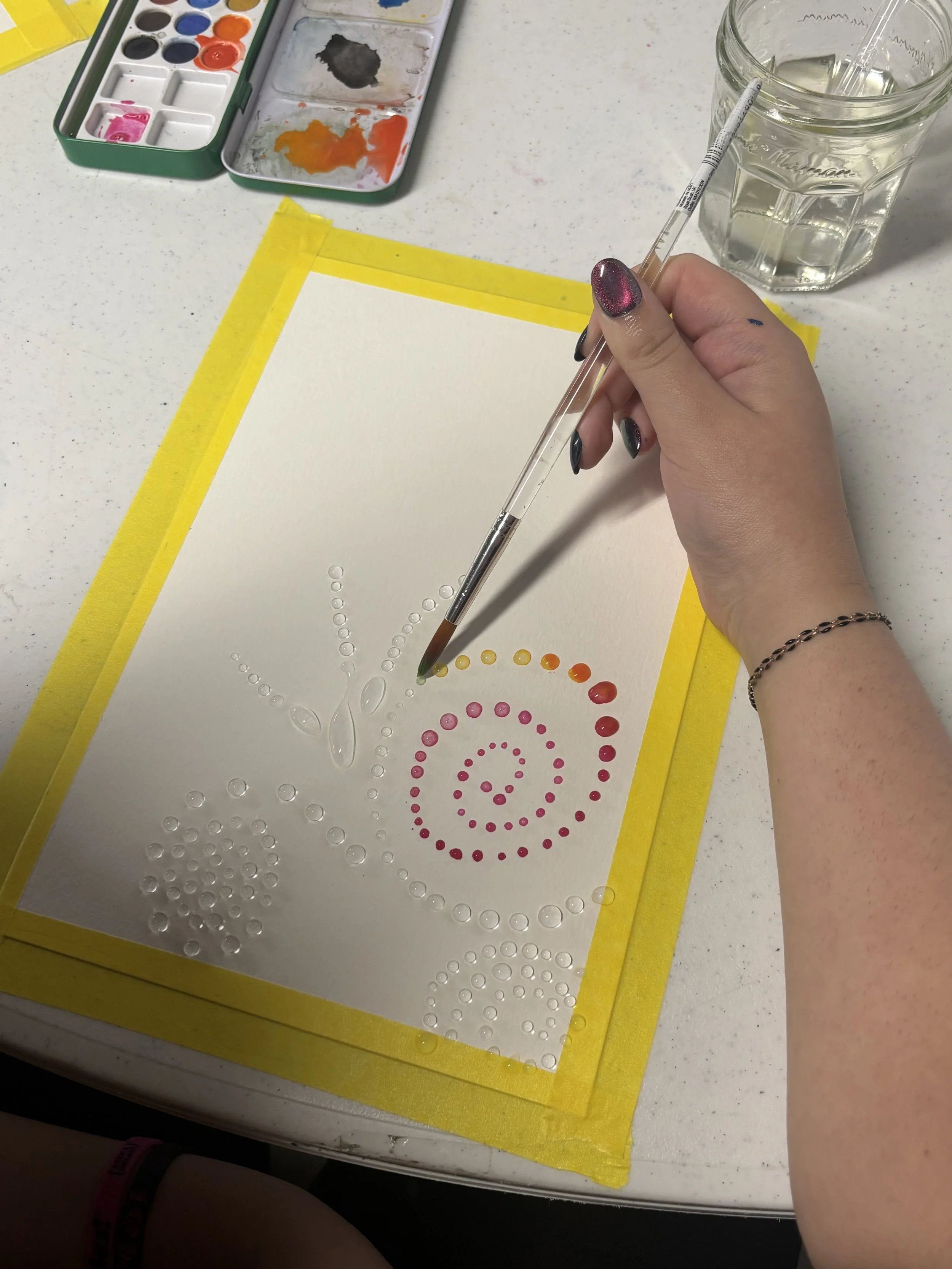 A person creates a dot mandala pattern on paper using watercolor paints and a pipette, with a palette of mixed colors and a glass of water nearby.