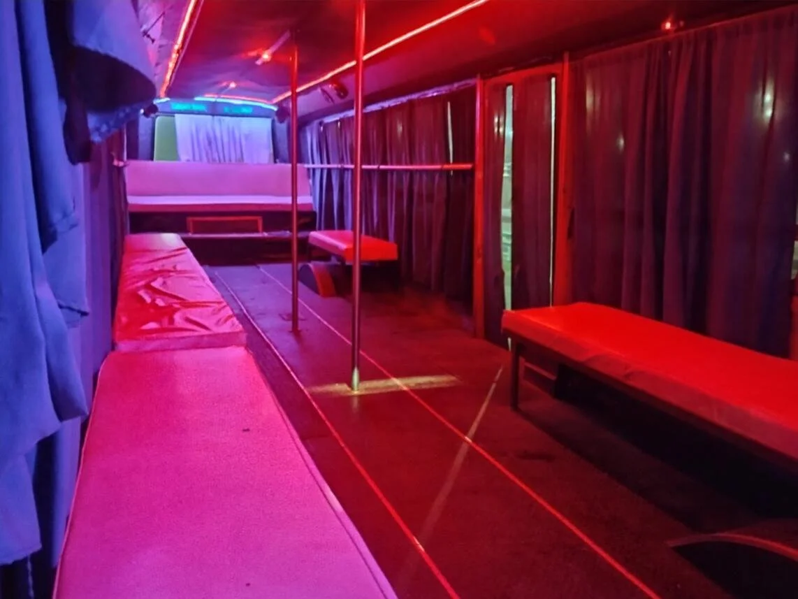 Interior de un autobús decorado con luces de neón rojas y azules, con cortinas oscuras, asientos rojos y barras metálicas para pasajeros.
