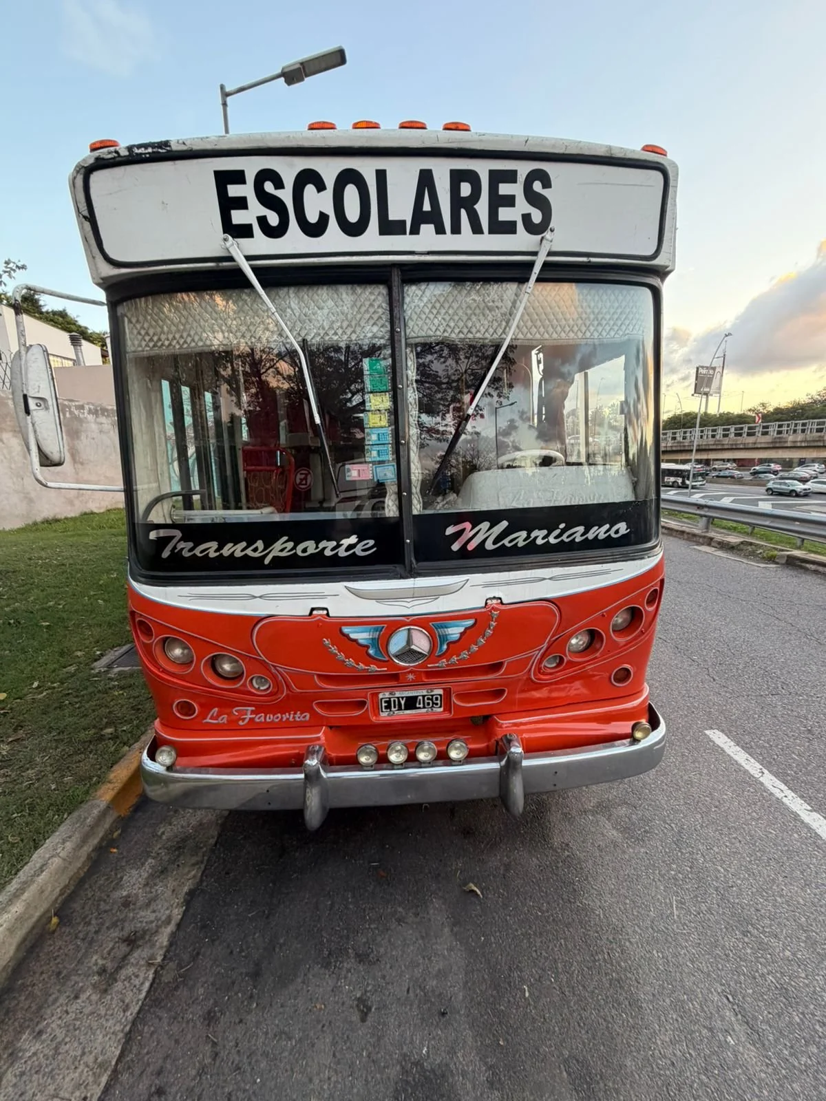 Furgoneta de transporte escolar de color rojo con letrero 'ESCOLARES' en la parte superior y la palabra en la parte frontal 'Transporte Mariano' y 'La Favorita', con un logo en el centro y luces en la parte delantera