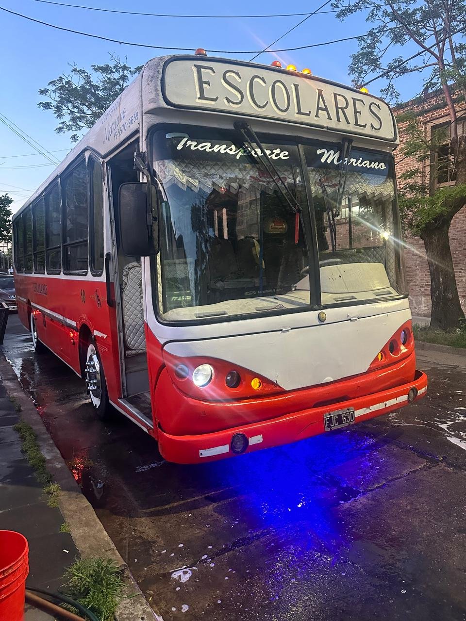 Un autobús escolar pintado de blanco y rojo con letrero en el parabrisas que dice 'ESCOLARES' y con faros delanteros encendidos, estacionado en la calle con un cielo despejado y árboles en el fondo.