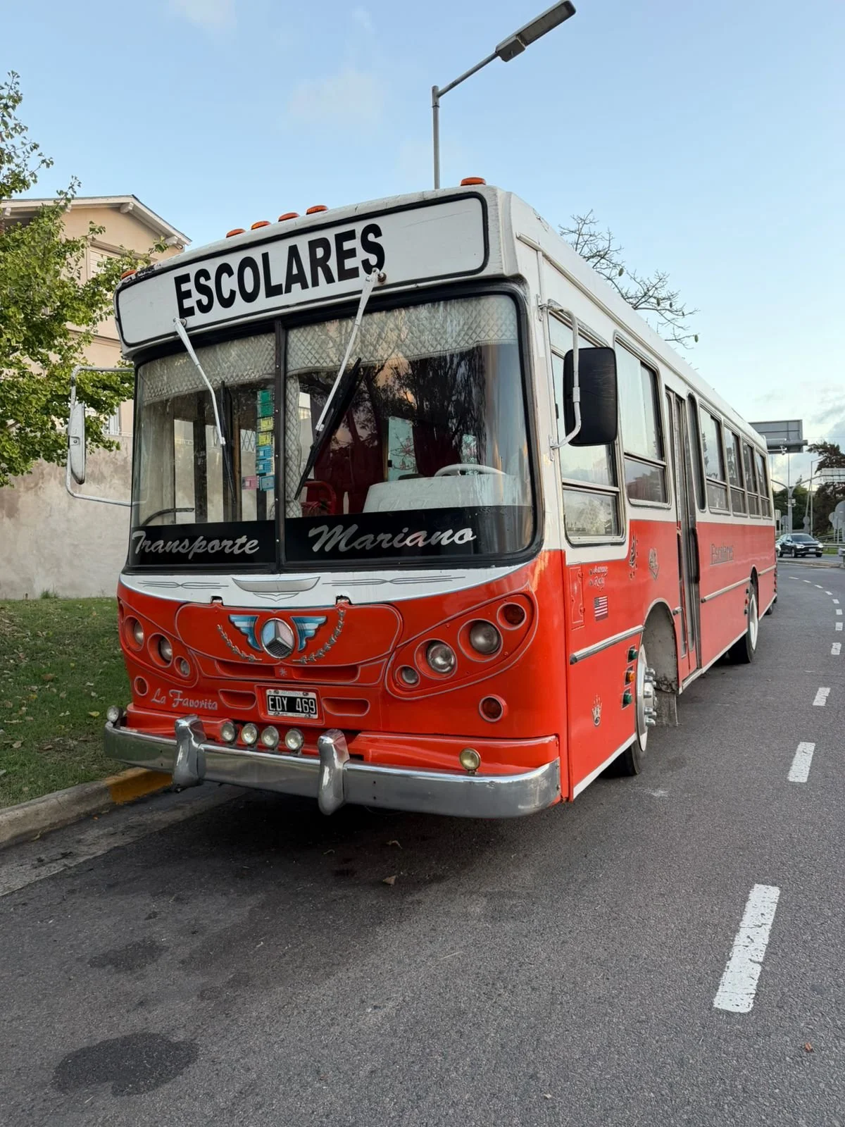 Autobús escolar rojo estacionado en la calle, con letrero que dice 'Escolares' en la parte superior y nombre 'Transporte Mariano' en el parabrisas.