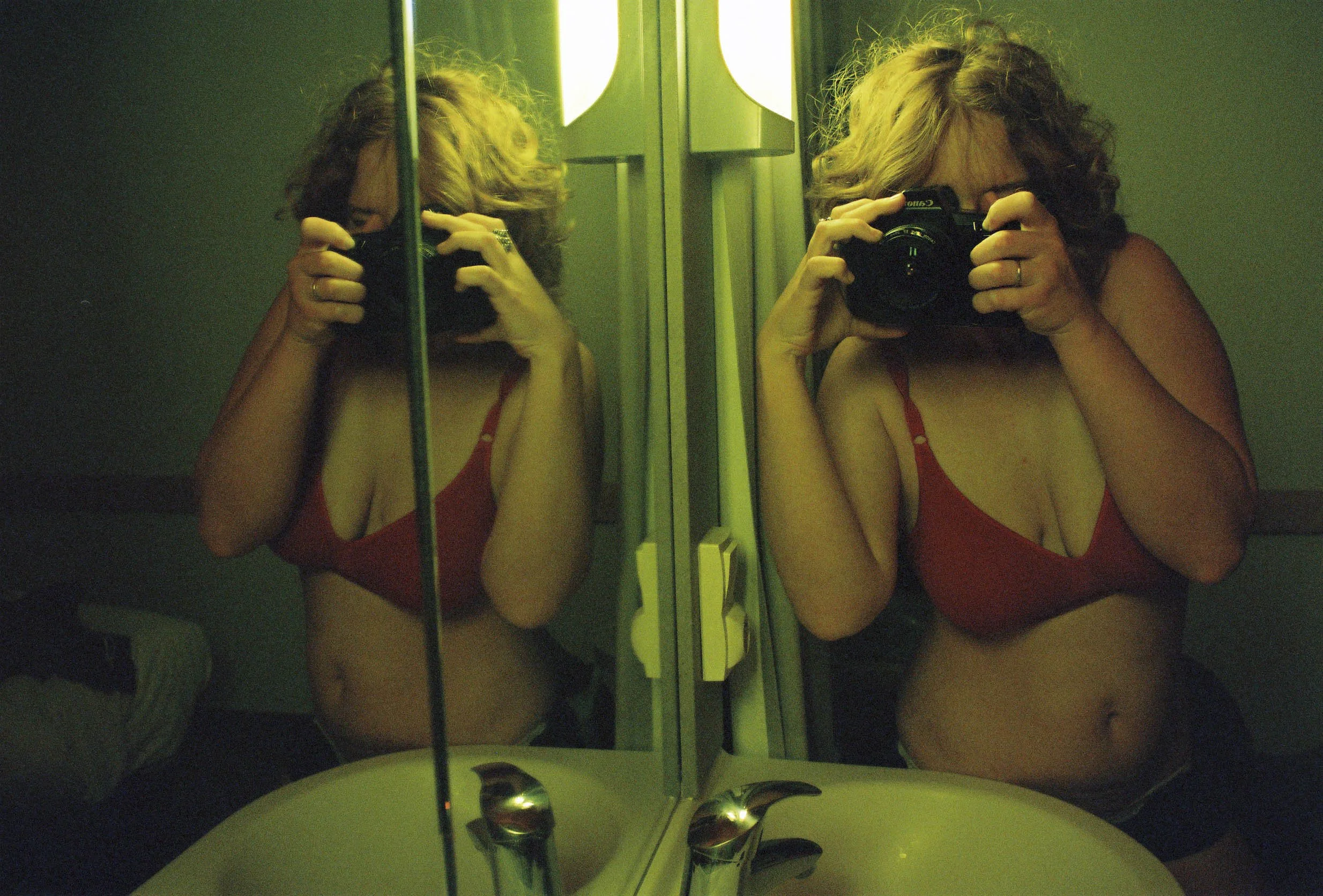 A woman with curly red hair taking a photo of herself in a mirror using a camera. She is wearing a red top, and the photo shows her reflection with green-colored lighting. Atmosphere is intimate and even mystic.
