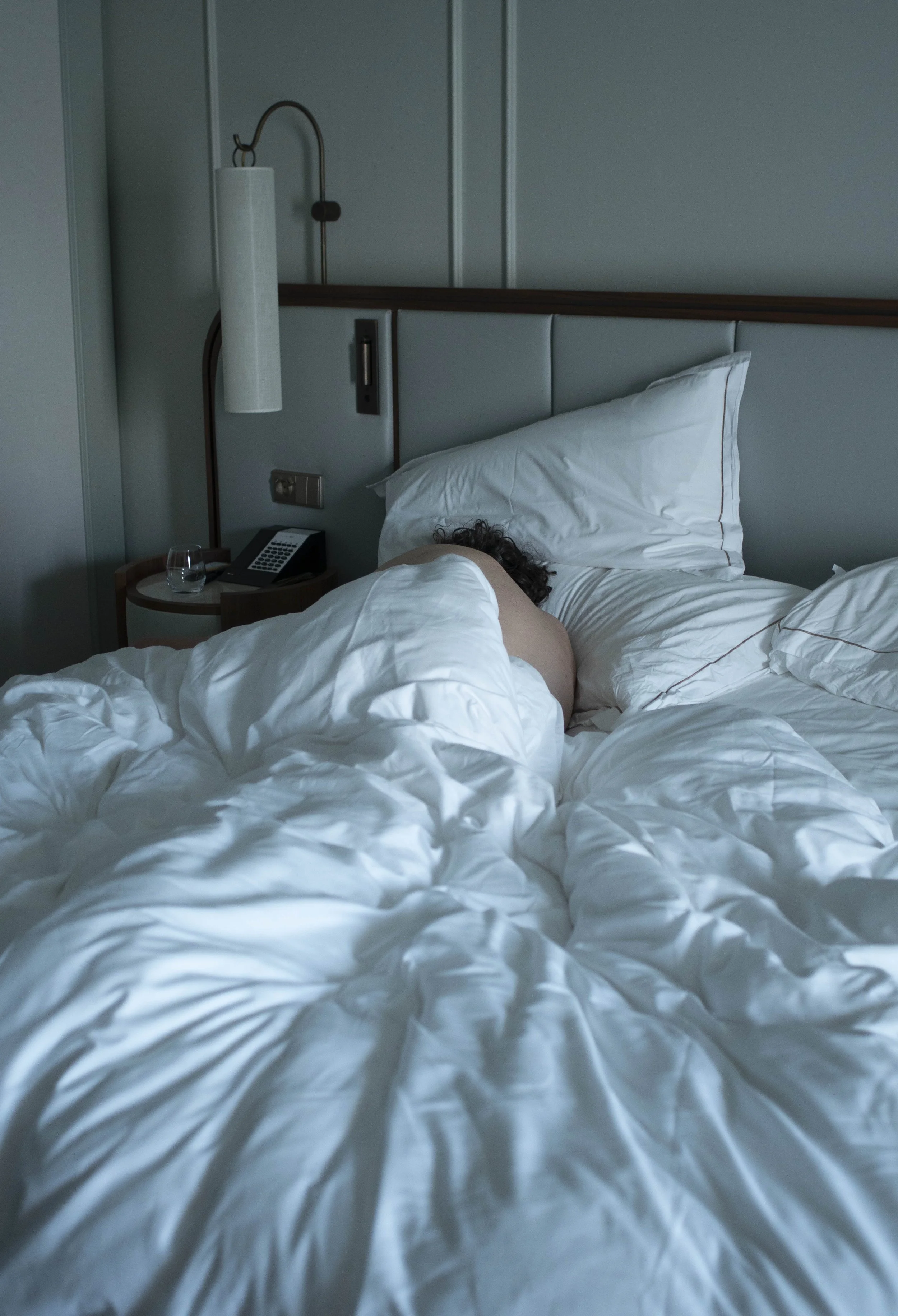 A person lying in bed with white bedding, head on a pillow, in a hotel room with a bedside table, phone, glass of water, reading lamp, and wall paneling.