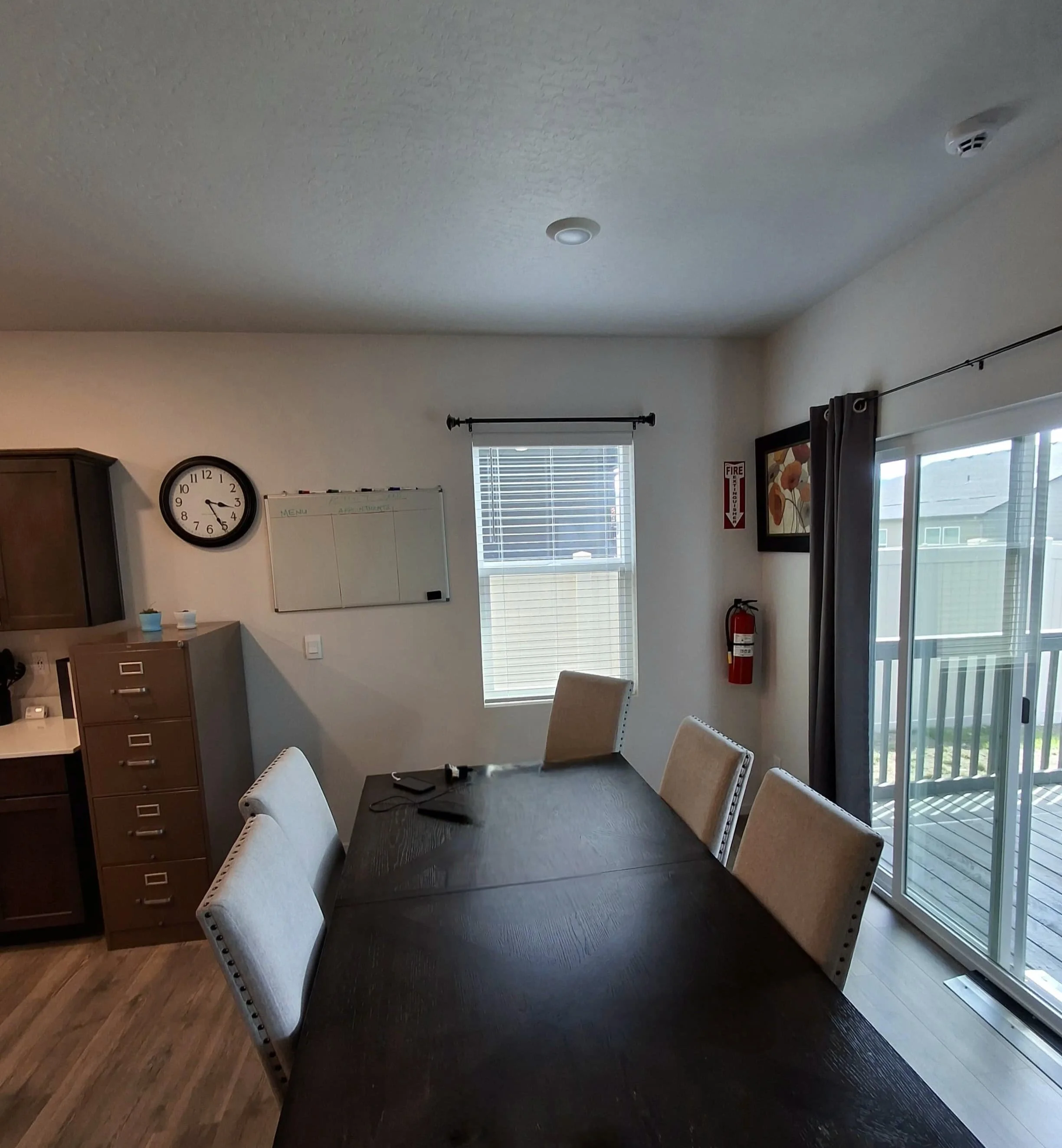 A dining room with a dark wooden table, white upholstered chairs, a clock, a whiteboard, two windows with blinds, and sliding glass doors leading to a balcony.