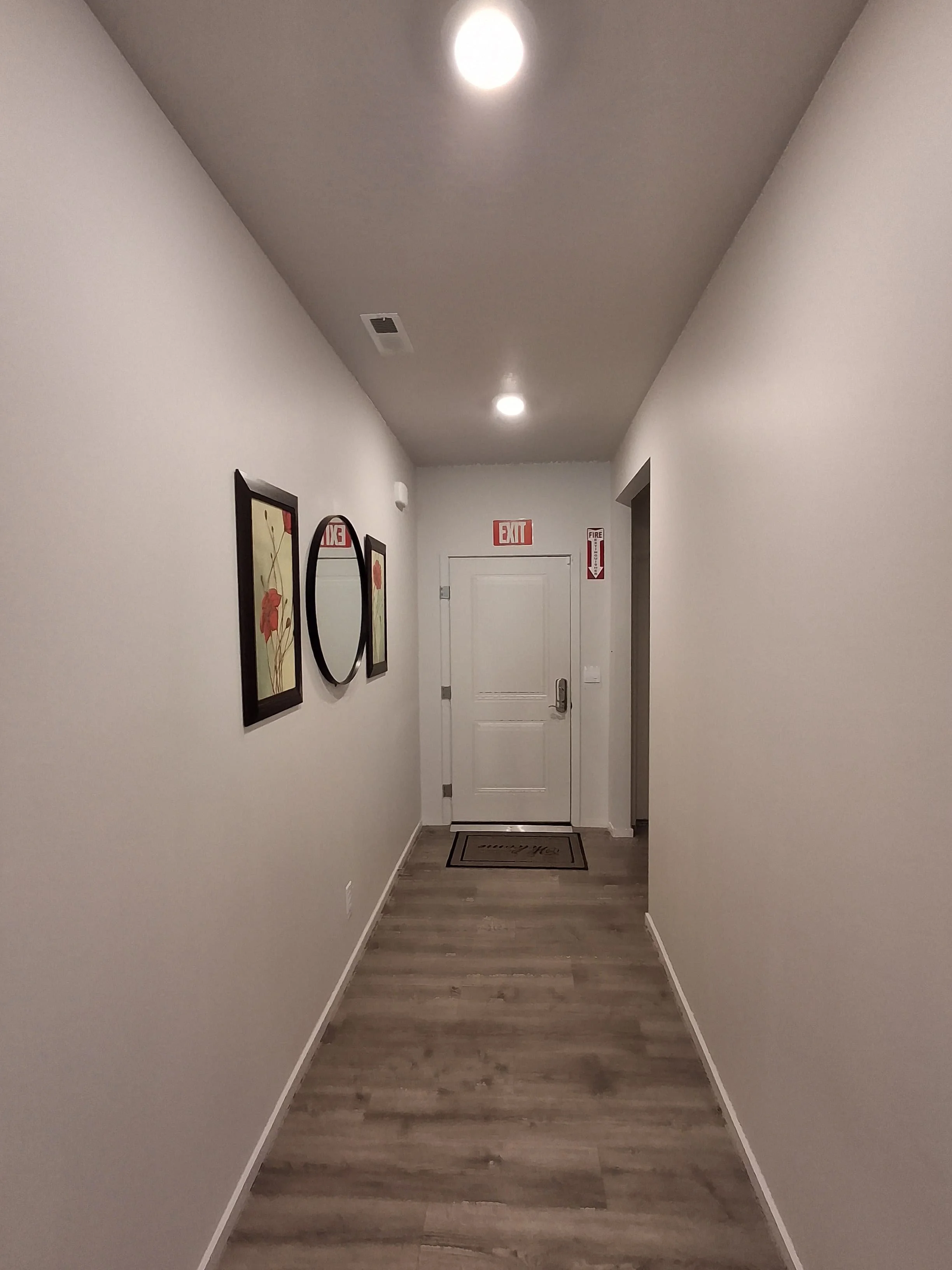 A hallway with white walls, wooden flooring, and framed floral artwork on one side. The end of the hallway has a white door with an electronic lock, and above it, emergency exit and fire safety signs. Recessed ceiling lights illuminate the space.