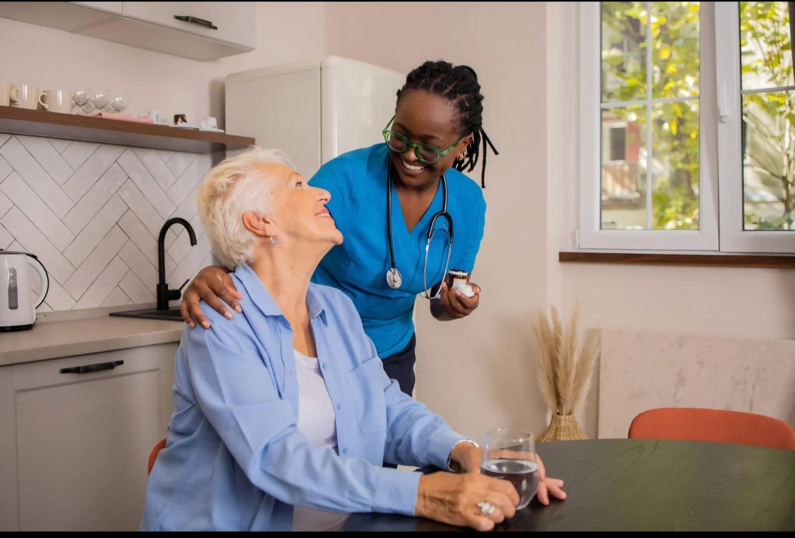 An elderly woman sitting at a table with a glass of water, talking happily with a healthcare worker who is standing next to her in a bright kitchen. The healthcare worker is smiling and has her hand on the woman's shoulder.