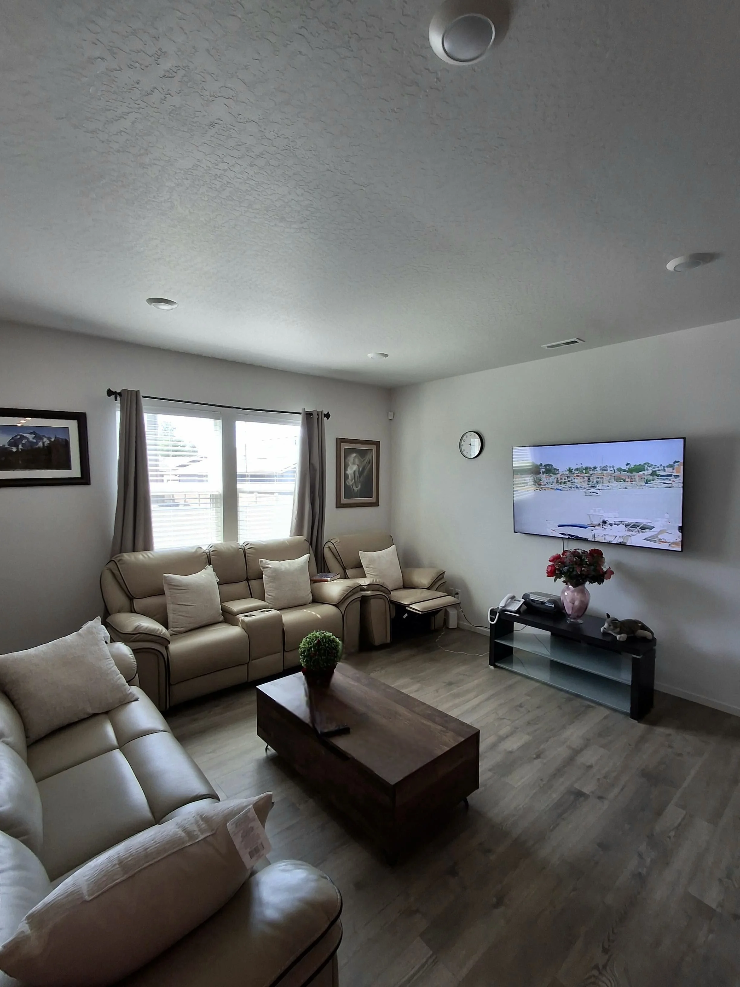 Living room with leather sofas, a wooden coffee table, a TV mounted on the wall, and framed artwork, with a window covered by curtains and a clock on the wall.