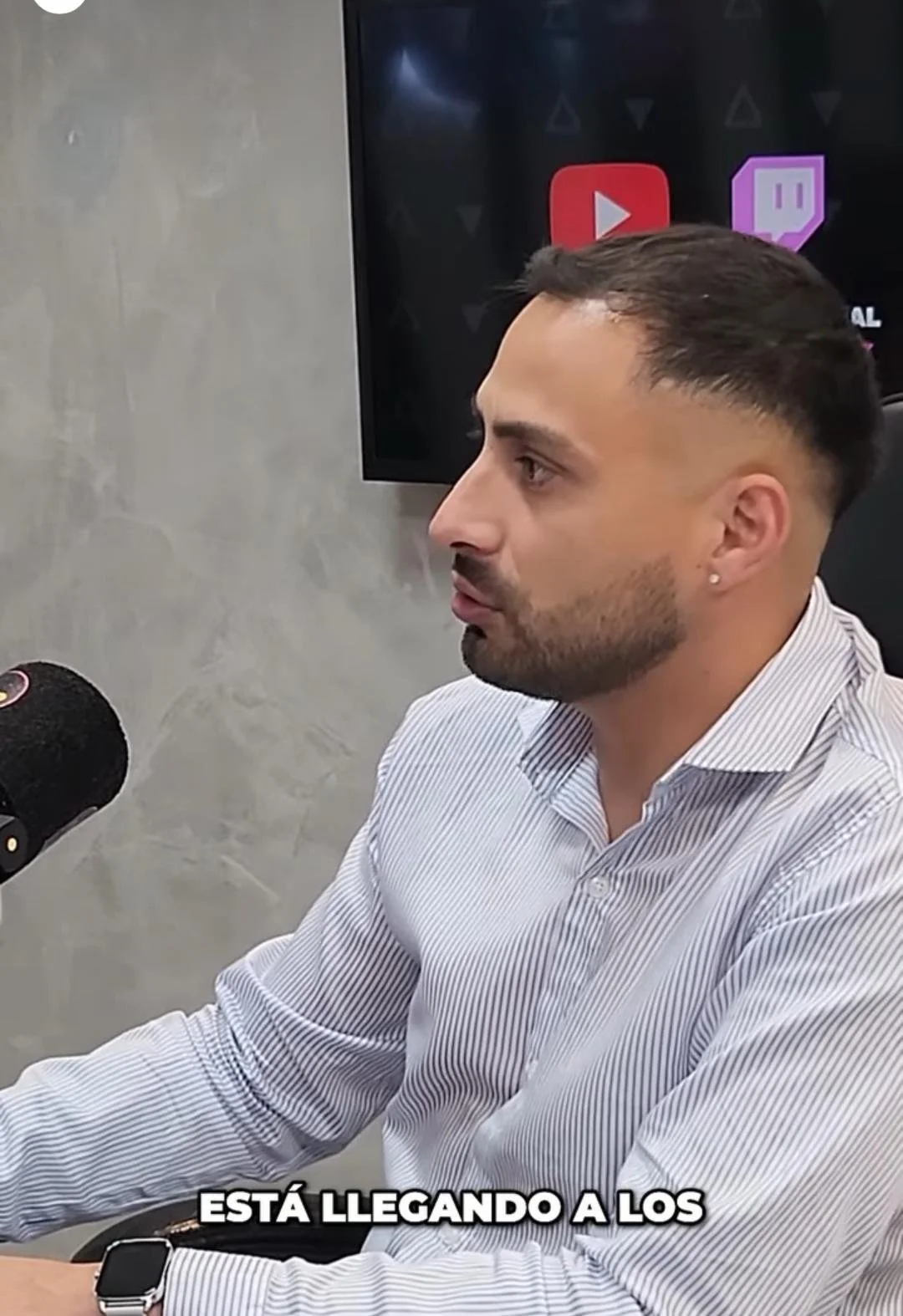 Hombre de perfil hablando en una entrevista, con teléfono en la mano, en un ambiente de oficina, en el fondo hay una pantalla con logotipos de YouTube y Twitch.