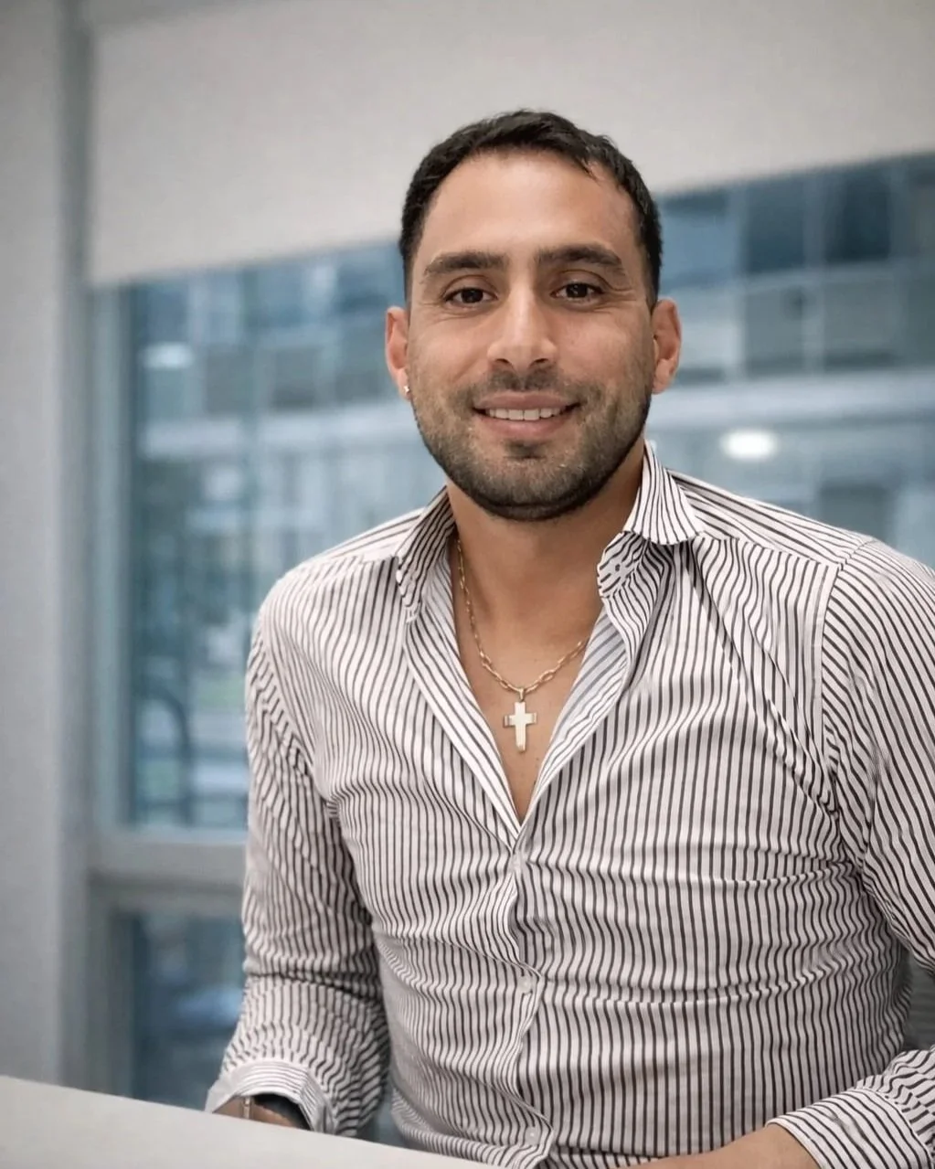 Hombre sonriendo en una oficina, lleva una camisa de rayas y un collar con cruz.