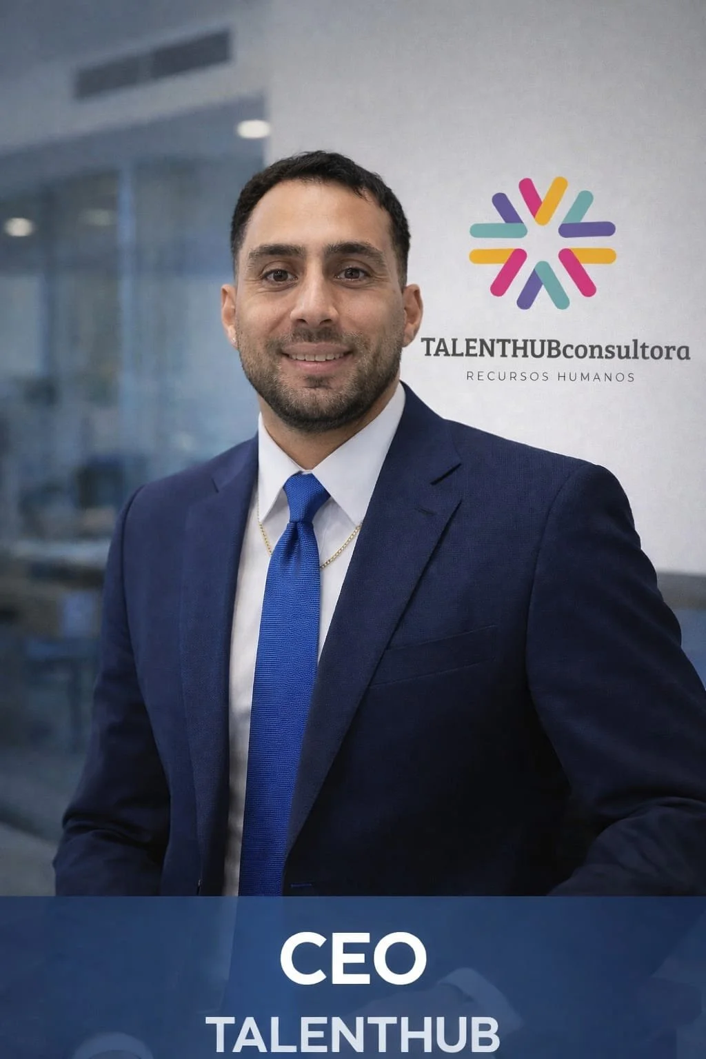 Hombre de negocios con traje azul y corbata, posando frente a un cartel de la empresa TALENTHUB consultora, que se dedica a recursos humanos, en un entorno de oficina moderna.