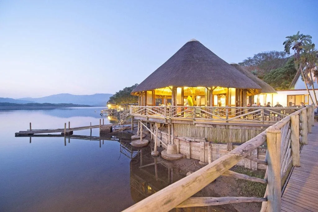 Lakeside restaurant with thatched roof, wooden deck, and boat dock, illuminated at dusk with calm water reflecting the structure.
