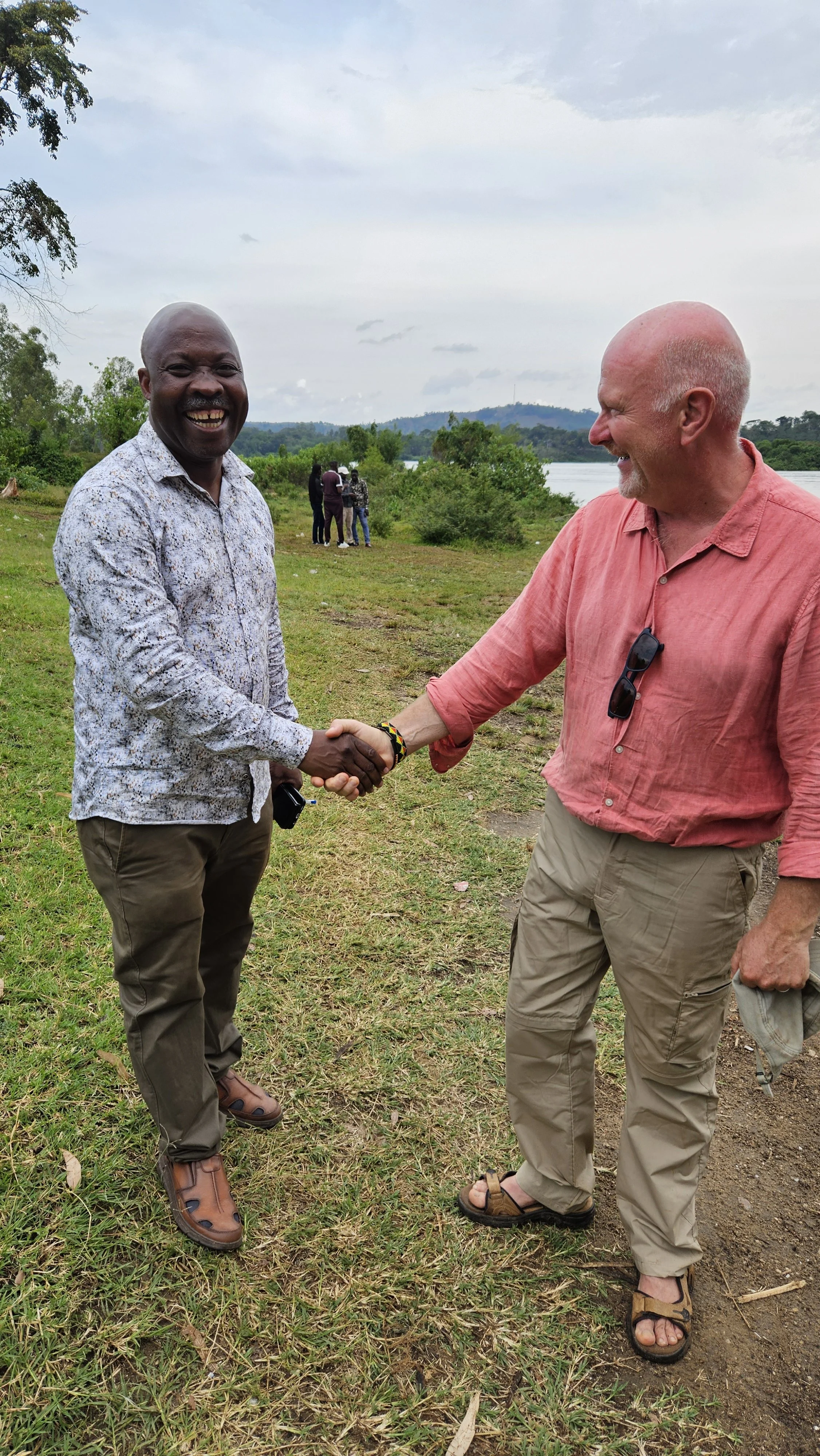 Two men are shaking hands outdoors near a body of water with trees and hills in the background. One man is wearing a patterned shirt and brown pants, the other is wearing a red shirt and khaki pants. They are smiling and appear to be greeting each ot
