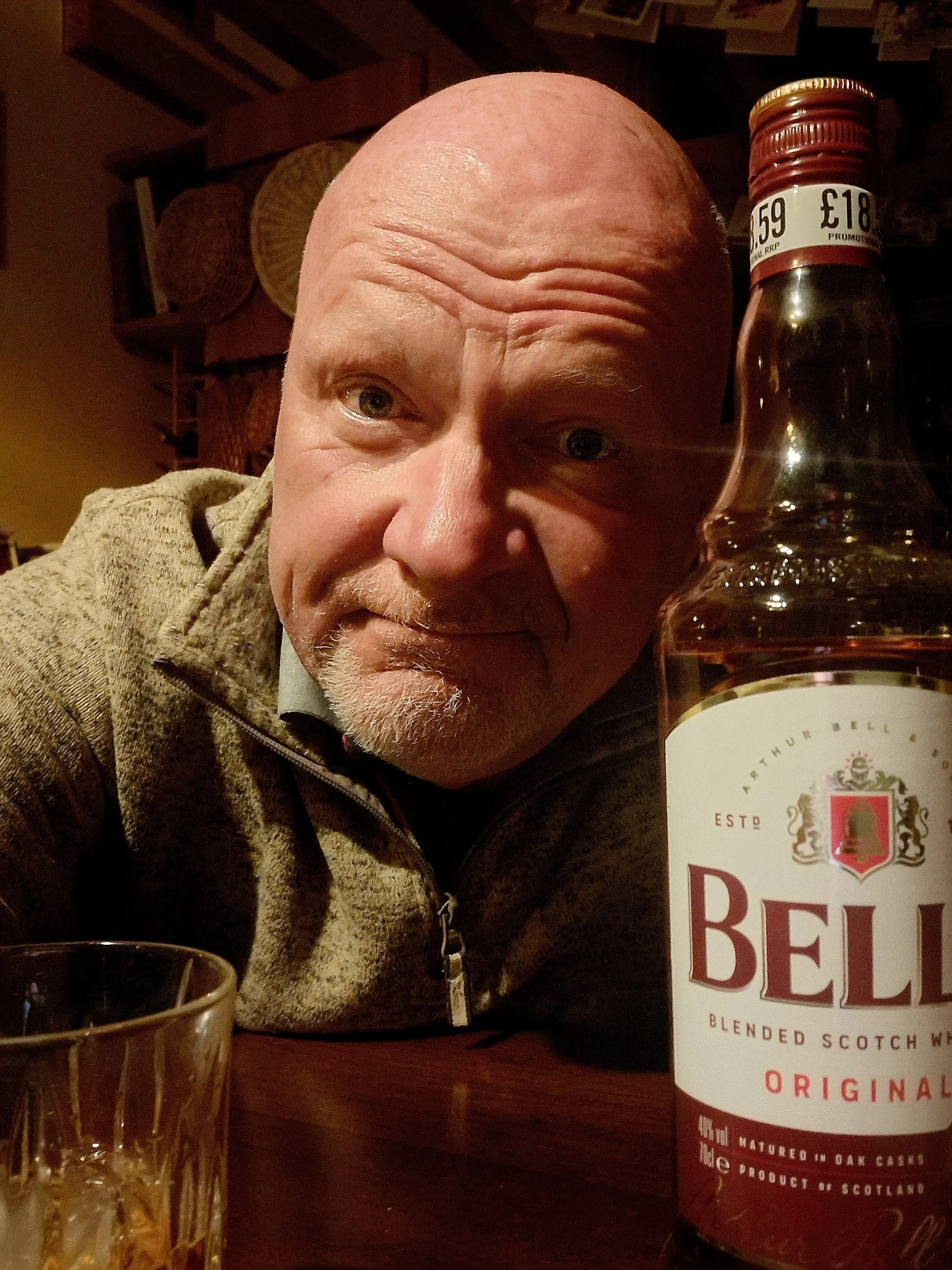 A close-up of a bald man with a beard, sitting at a table with a glass of dark beverage in front of him, and a bottle of blended Scotch whisky labeled "The Famous Grouse" on his right side.