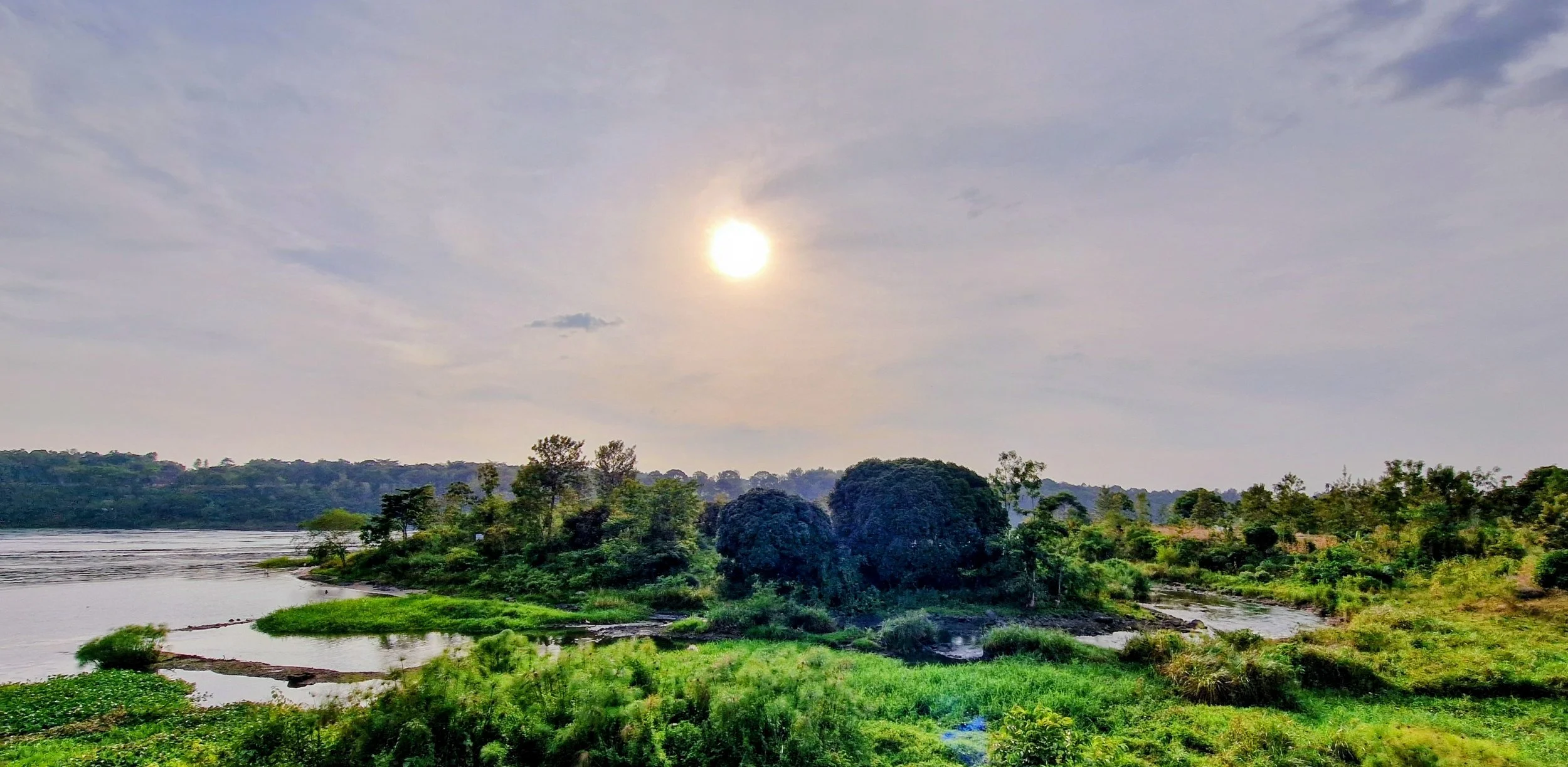 A scenic landscape of the Nile river with lush green trees along its banks, under a cloudy sky with the sun peeking through.