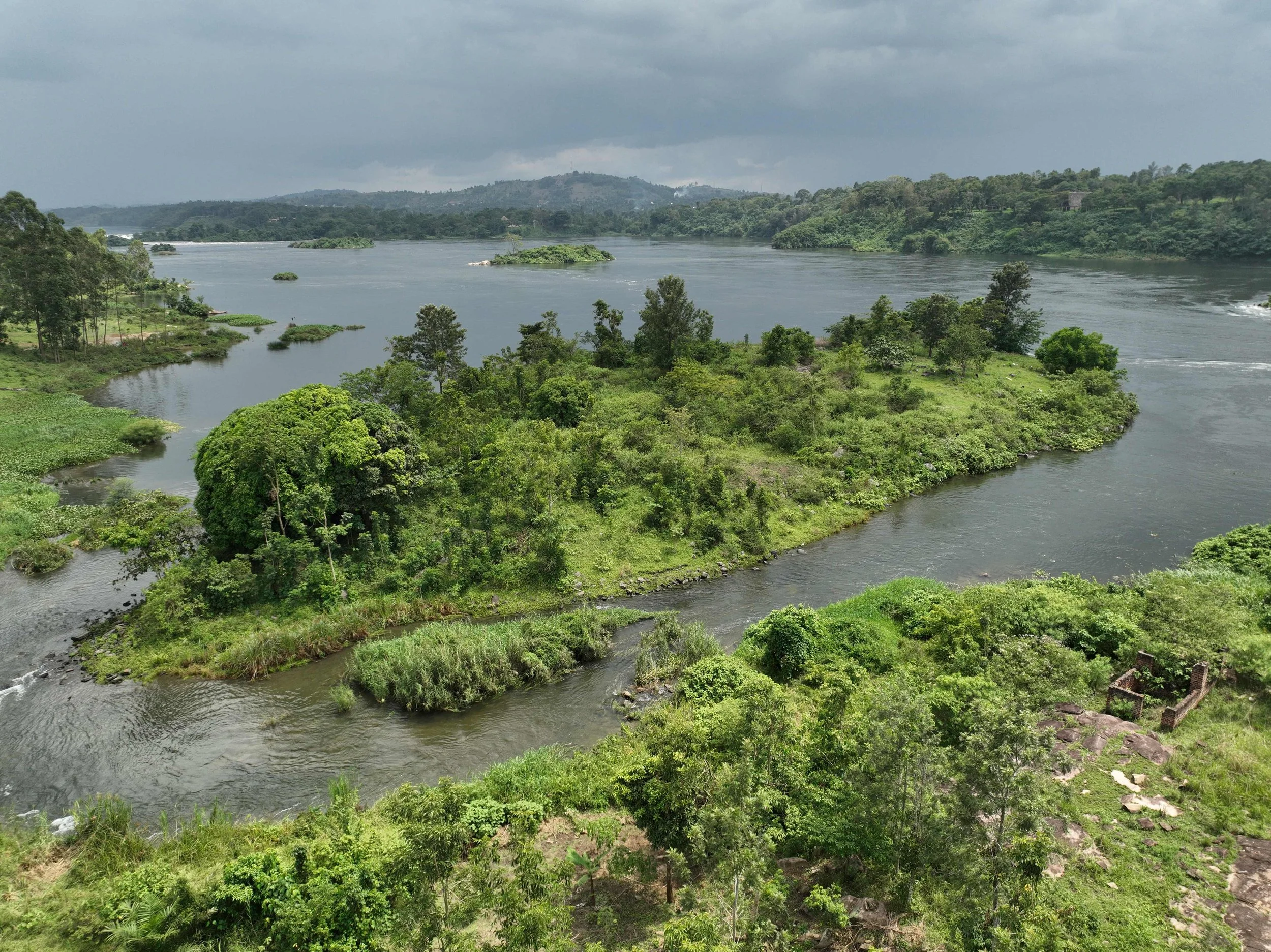 A large river flowing through lush green landscape with small islands and hilly terrain in the distance, under a cloudy sky.