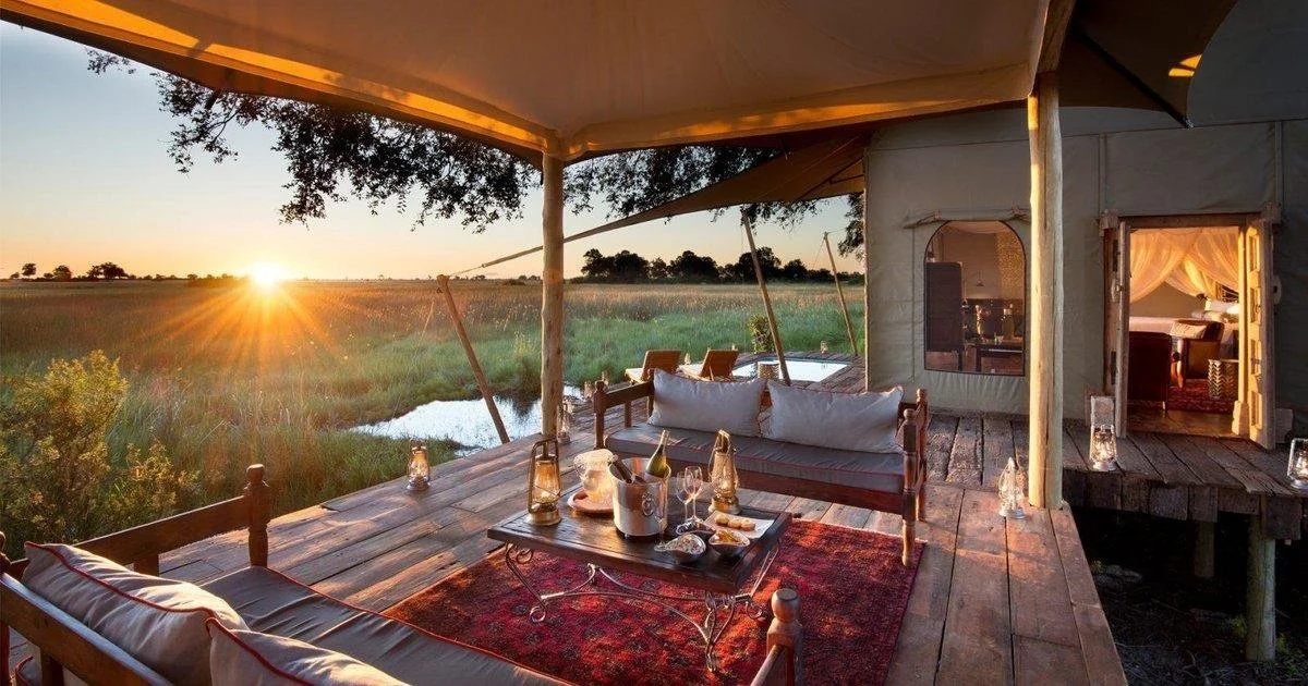 Outdoor patio at sunset with wooden furniture, lanterns, and a view of the open fields and water.